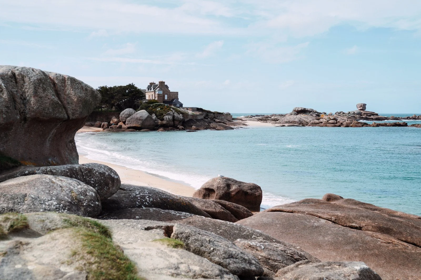 Plage rocheuse avec une maison en arrière-plan, eau calme, rochers au premier plan, ciel en partie nuageux.