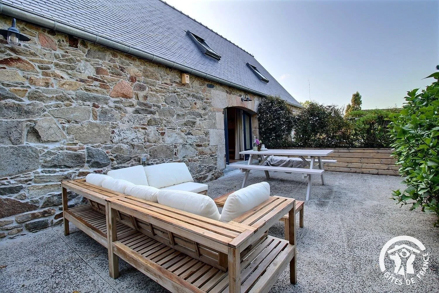 Terrasse extérieure avec canapé en bois et coussins blancs, table en bois avec une bouteille et des fleurs, murs en pierre et végétation encadrant l'espace, ciel clair.