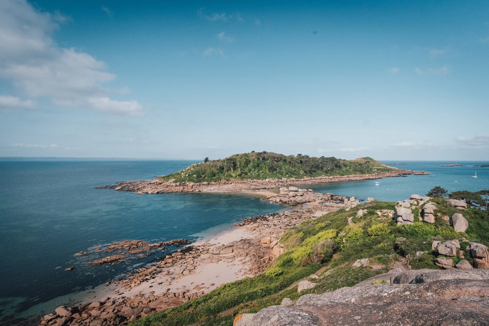 Vue d'une petite île rocheuse couverte de végétation, entourée d'eau bleue, avec un ciel clair et quelques voiliers au loin.