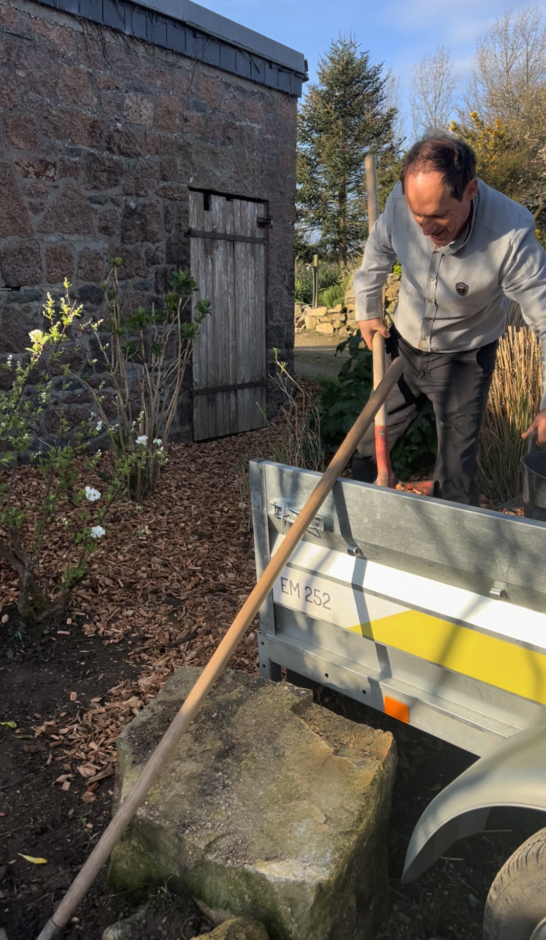 Un homme souriant utilise une pelle pour déplacer des matériaux dans une remorque en métal attachée à un véhicule, dans un jardin avec une petite plantation de fleurs et une vieille cabane en pierre en arrière-plan.