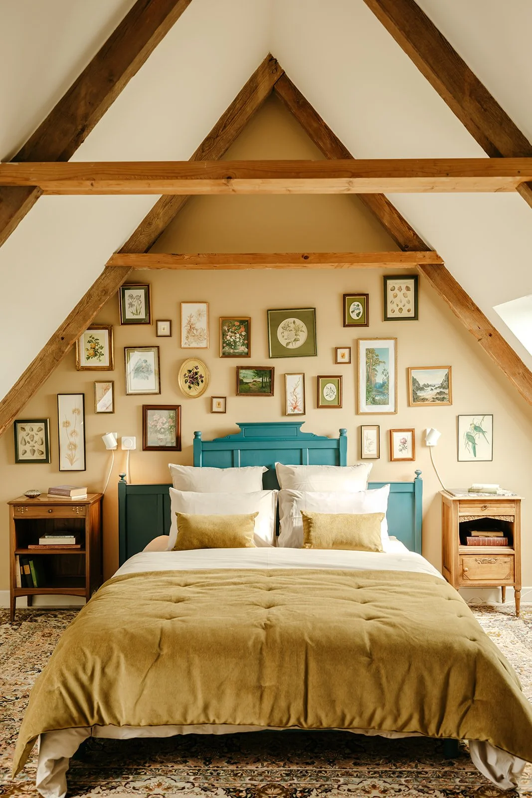 Chambre avec un lit à baldaquin turquoise, murs décorés de nombreux cadres avec des illustrations botaniques, deux tables de chevet en bois et des lampes murales, tapis persan, murs en angle avec poutres en bois apparentes.