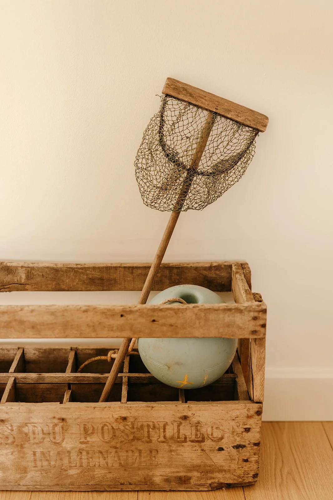 Une caisse à bouteilles en bois de brocante contenant une bouée et une épuisette en bois, le tout posé contre un mur beige.