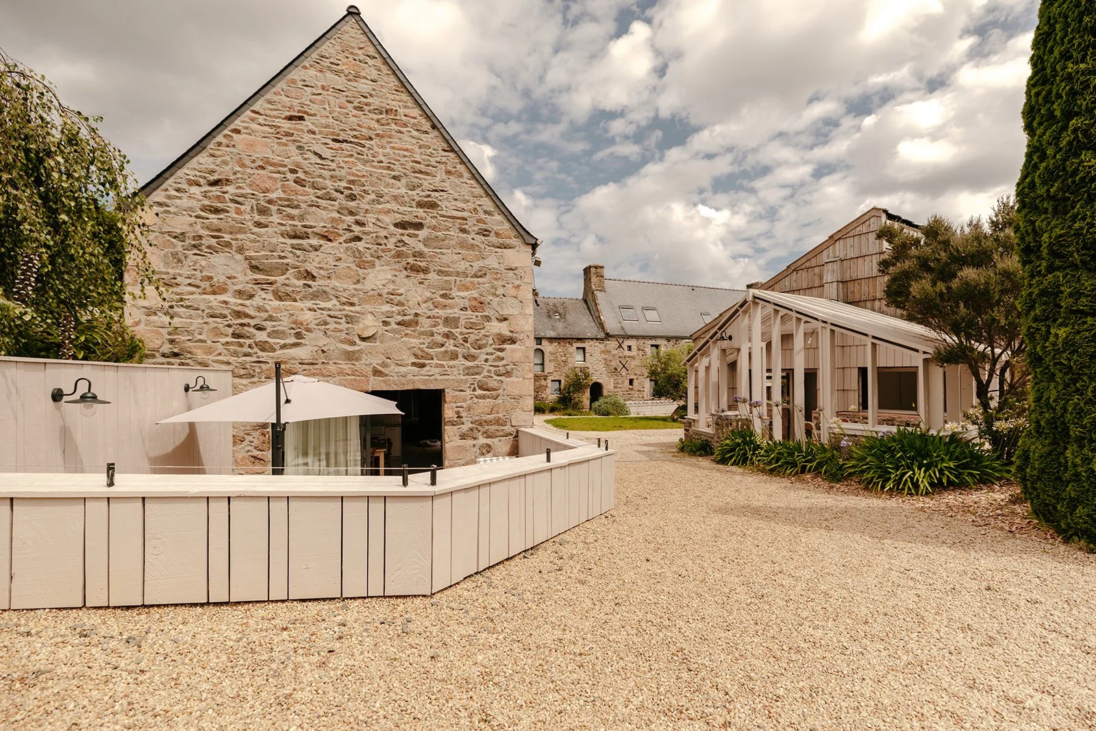 Cour extérieure d'une ferme en pierre rénovée avec une terrasse, un parasol blanc, et un bâtiment en bois, entourée de végétation et un chemin en gravier.