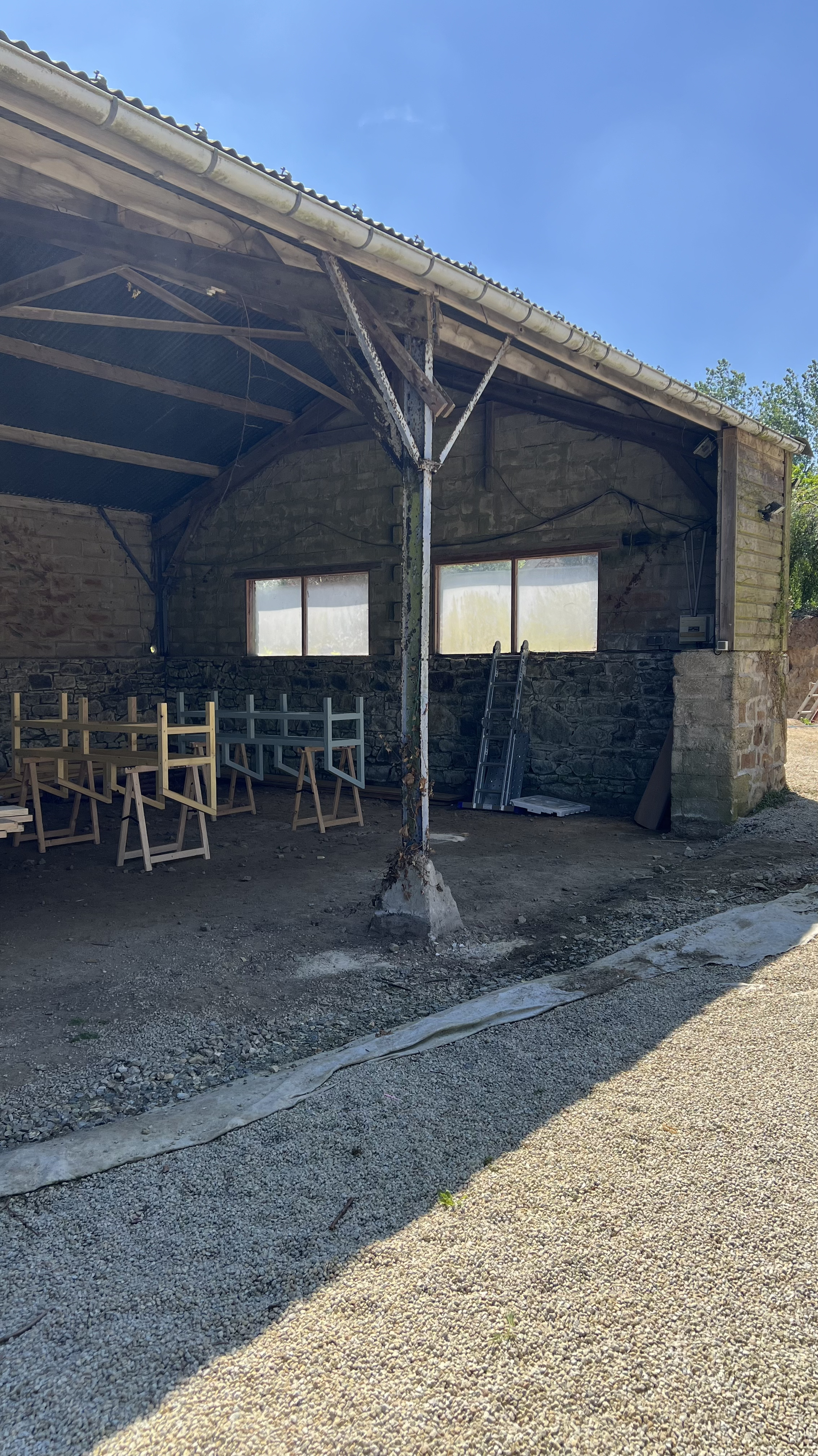 Une structure en construction ou en rénovation avec des murs en pierres, des fenêtres et une toiture partiellement installée. Il y a des échelles, des tréteaux en bois et des outils dispersés à l'intérieur.