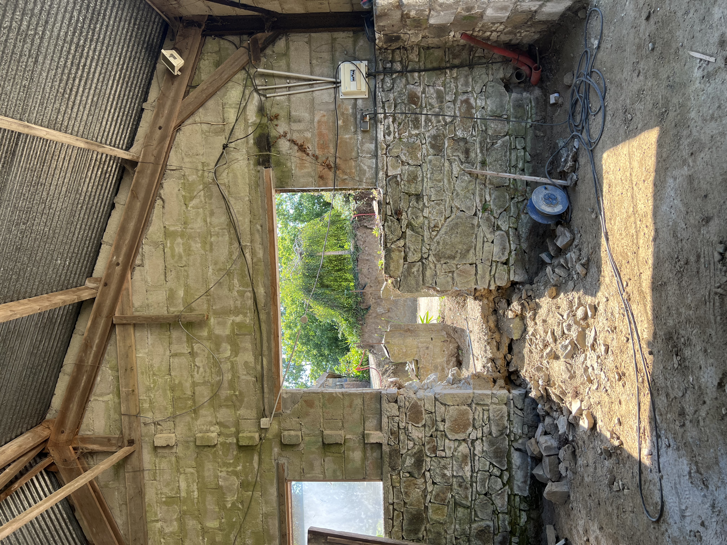 Une ancienne pièce de pierre en travaux, avec des murs en pierre, une ouverture, des outils électriques, et des pierres au sol.