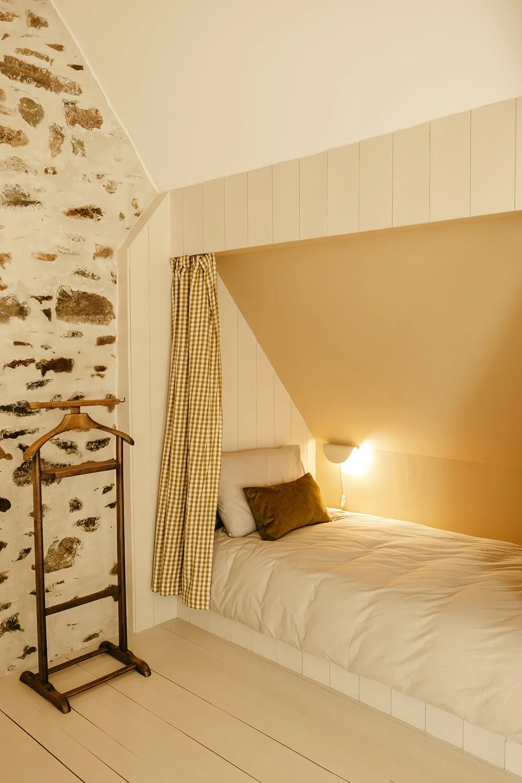 Chambre Padine du Clos des Sentiles à Perros-Guirec avec un lit simple sous pente, mur en pierre sur le côté gauche, draps blancs, coussin vert, lampe murale allumée, rideau vichy à carreaux jaune et blanc.