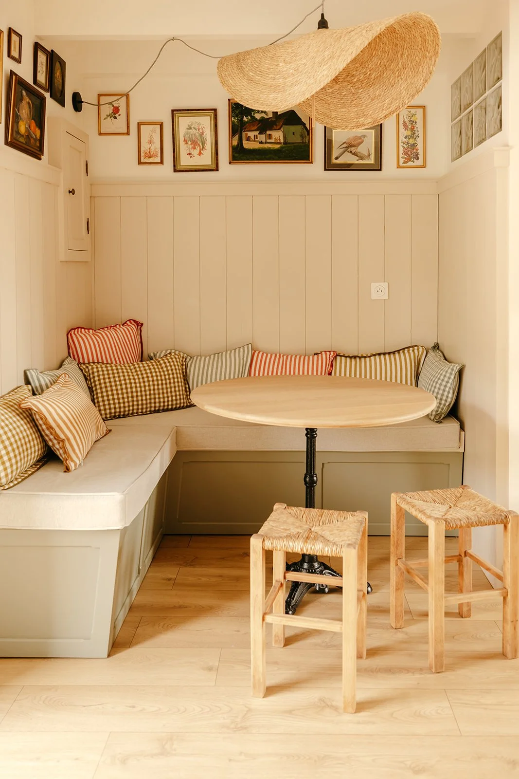 Coin repas avec un coin banquette rembourrée, un mur blanc avec des tableaux ornementaux, un lampadaire en osier et deux tabourets en bois avec le dessus en paille