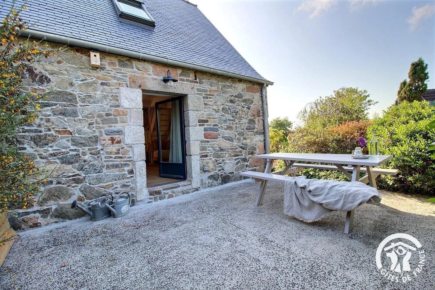 Terrasse extérieure de la chambre Padine au Clos des Sentiles avec table en bois, plantes en pot, et bâtiment en pierre avec porte-fenêtre, entourée de végétation.