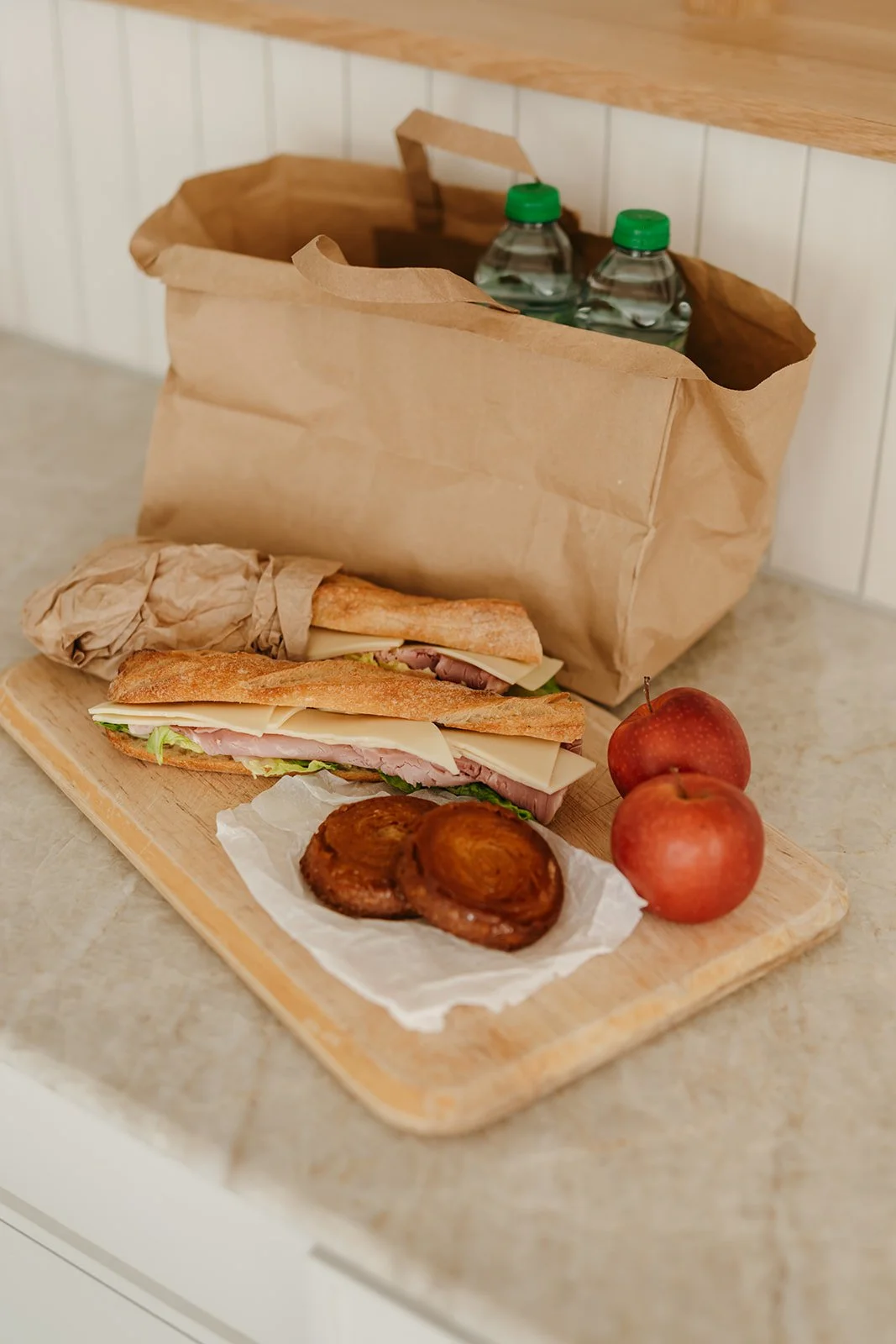 Un plateau en bois avec deux sandwichs au pain baguette, un croissant, deux pommes et deux petits pains aux raisins, une grande bouteille d'eau, et un sac en papier marron dans un environnement intérieur.