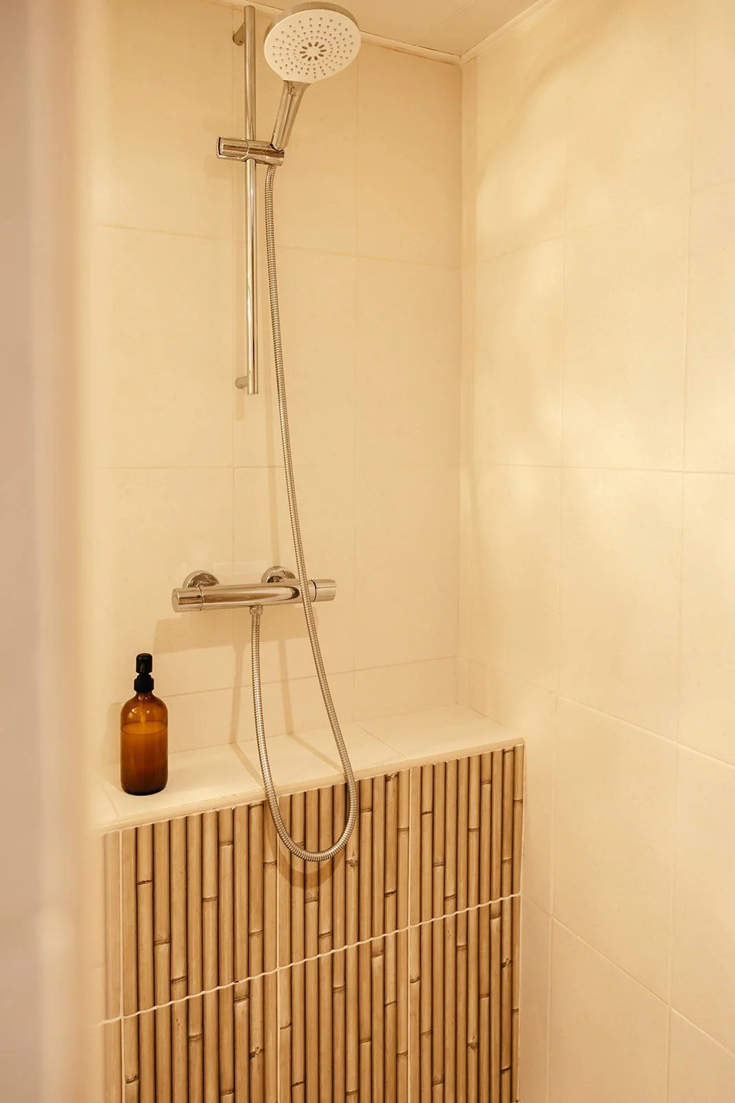Douche italienne avec pomme de douche ronde, une bouteille en verre ambré avec gel douche et des murs en carrelage beige.