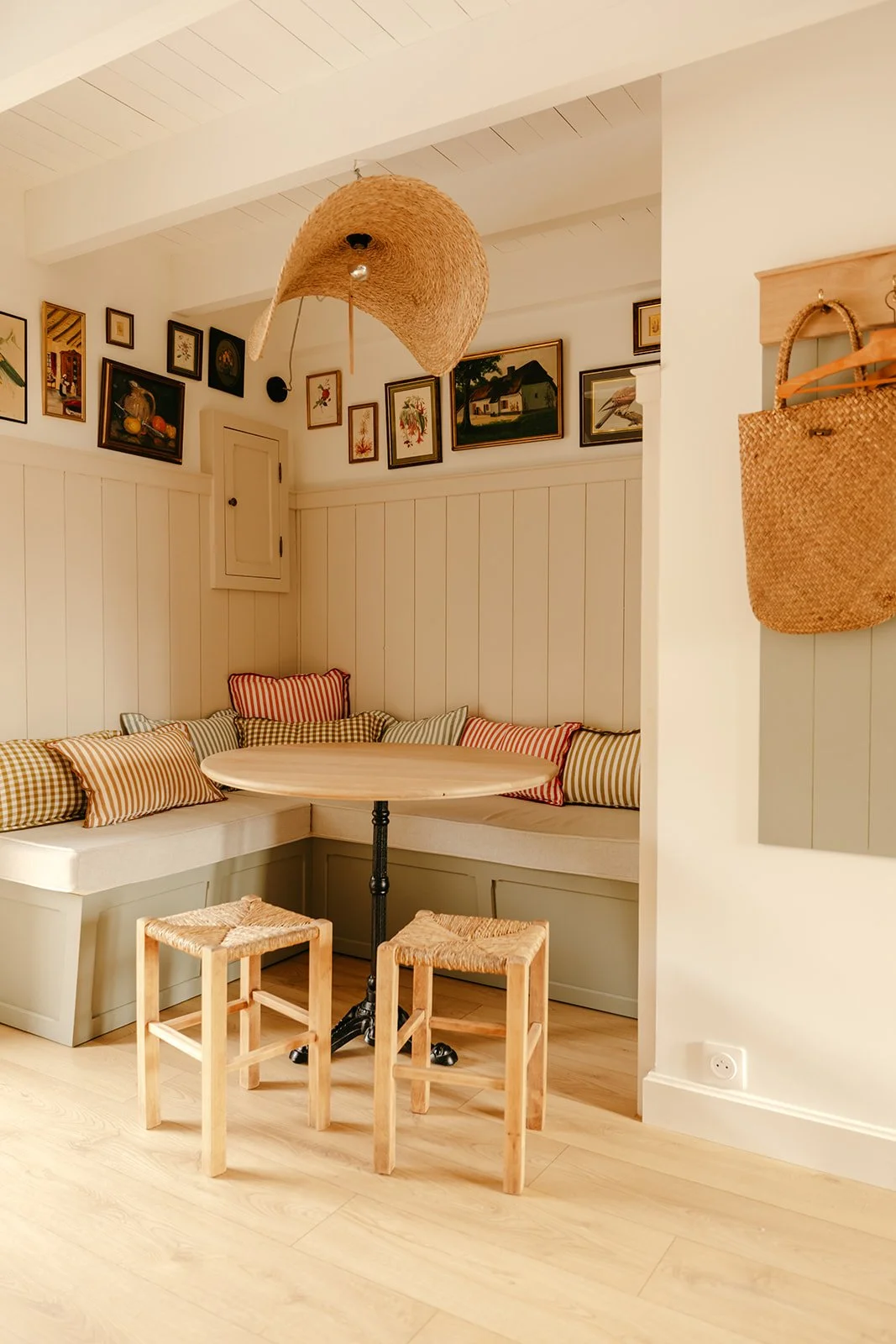 Coin dîner de la Chambre Padine du Clos des Sentiles avec une banquette rembourrée ornée de coussins vichy, une table ronde en bois, deux tabourets en bois, une galerie de cadres avec des œuvres d'art et un panier en osier suspendu à un mur.