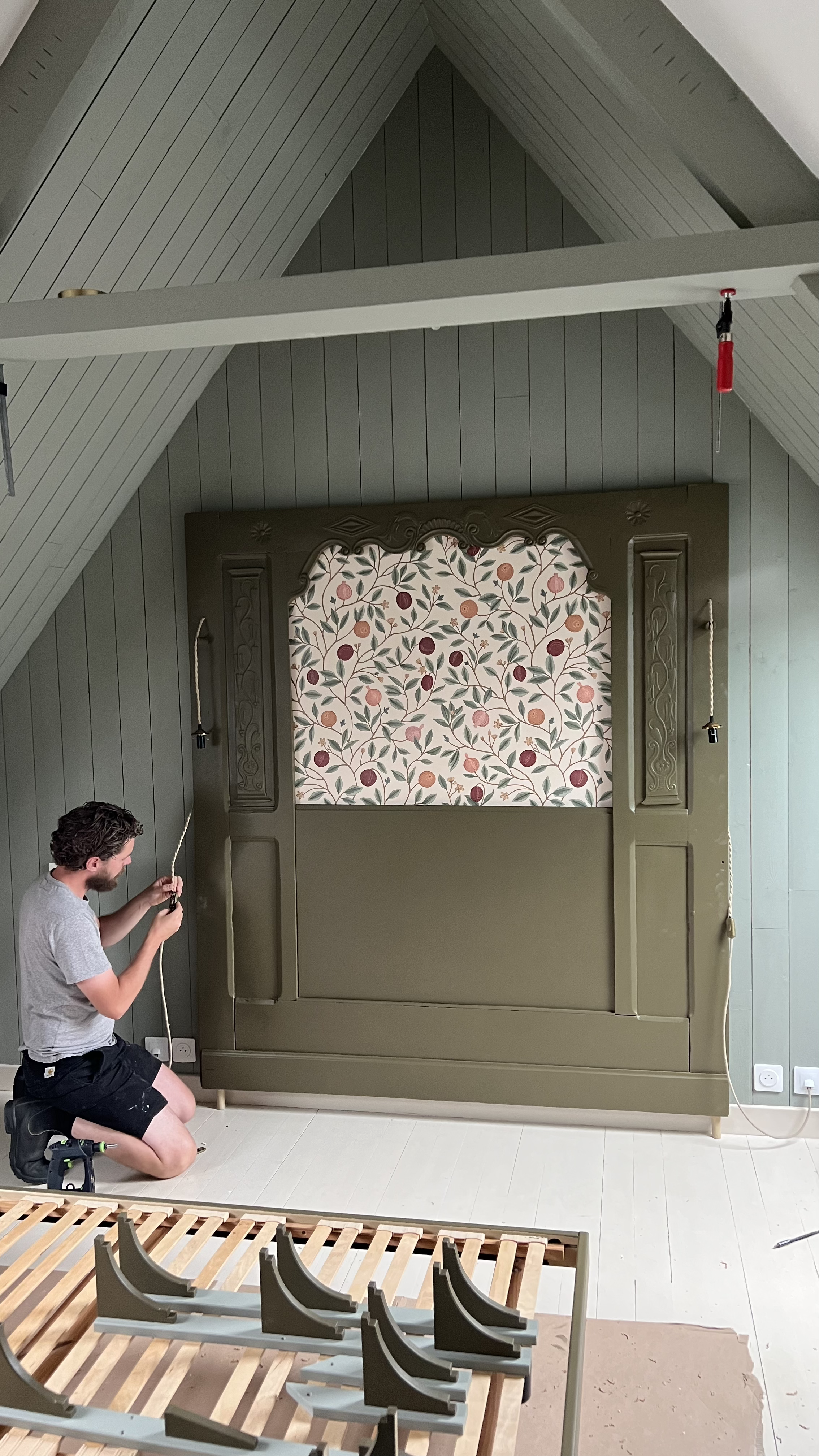 Une personne réassemble un lit dans une chambre, avec une tête de lit murale décorée de tissu floral. Il y a des pièces de lit en bois à l'avant et des outils de montage au sol.