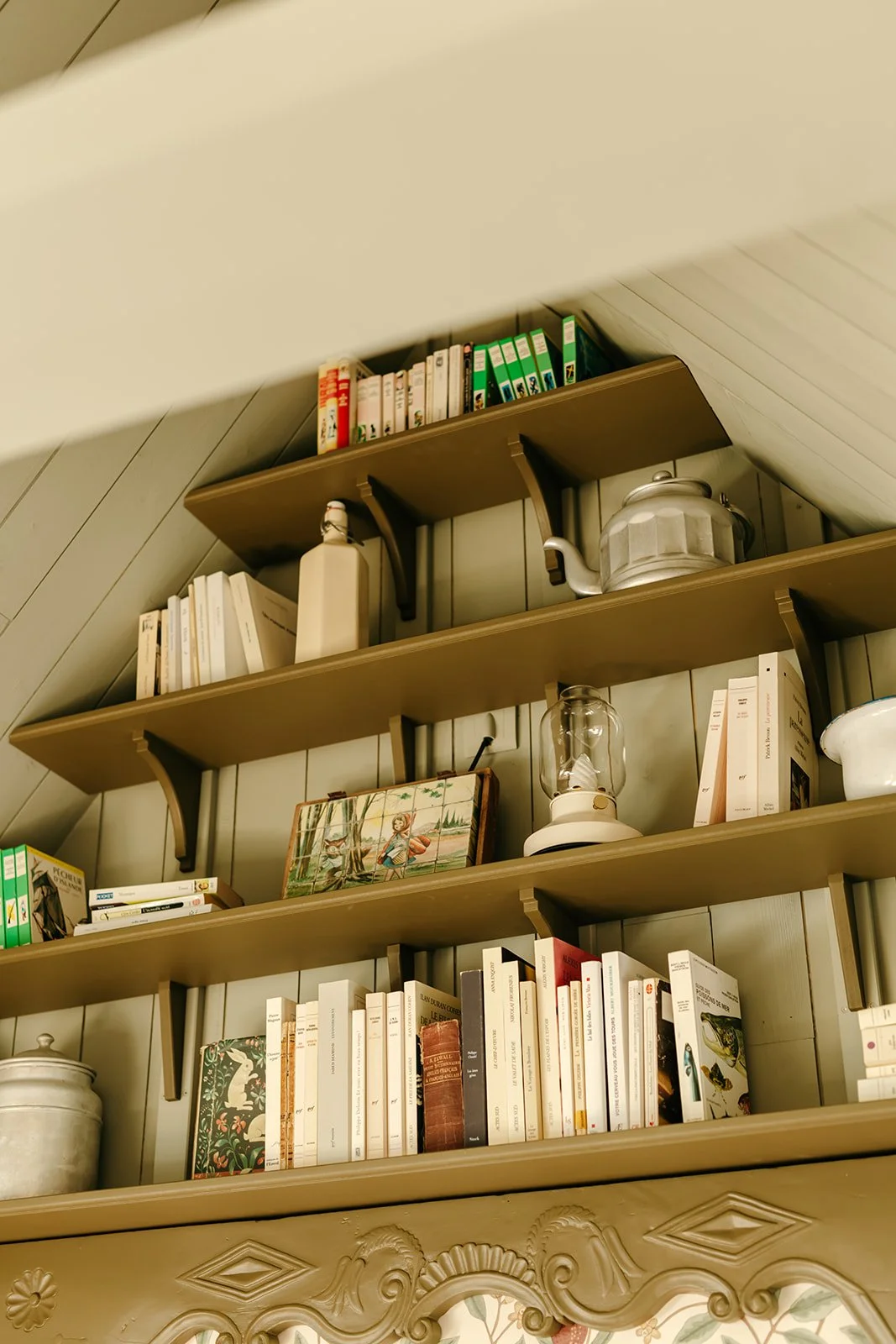 Étagères en bois peint, remplies de livres variés, une théière en métal, une lampe à huile, une bouteille en verre, et une peinture de paysage, dans une décoration de style vintage.