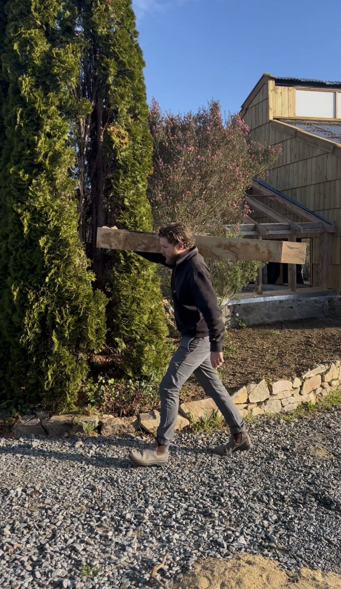 Un homme portant une grande poutre en chêne marche sur un sentier en gravier. Il traverse un jardin avec des plantes verts et un arbre en fleurs, avec un bâtiment en bois en arrière-plan.
