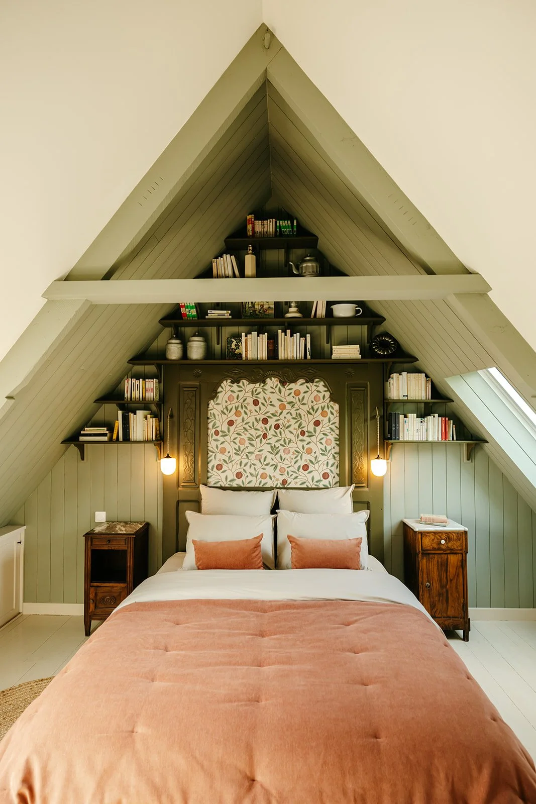 Chambre Dulse avec une tête de lit en bois, des étagères remplies de livres, un motif floral derrière la tête de lit, deux tables de nuit en bois de chaque côté, et un grand lit avec une couverture rose et des coussins roses.