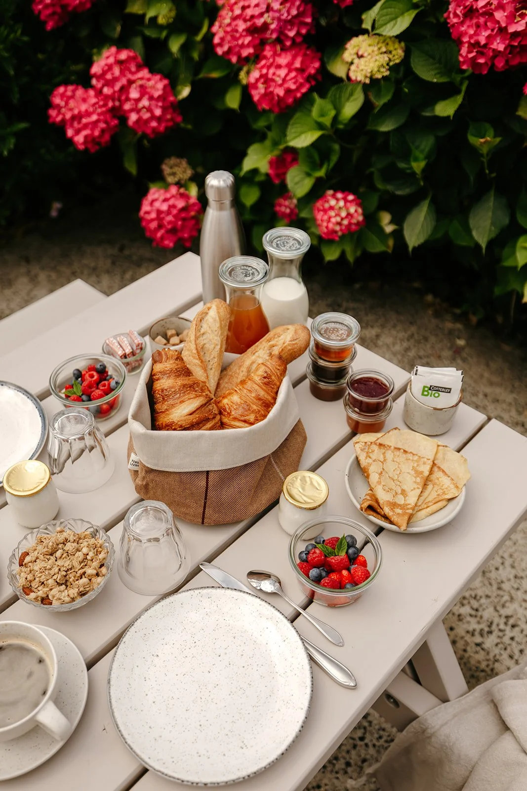 Petit déjeuner estival avec croissants, crêpes, fruits rouges, yaourts, confitures, miel, lait, eau, et café, sur une table en bois beige entourée d'hortensias roses et vertes.