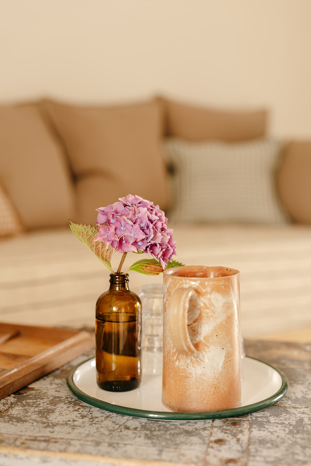 Une petite bouteille en verre marron contenant une branche avec une fleur de hortensia rose et des feuilles vertes, placée sur un plateau avec une tasse en céramique beige et un verre en arrière-plan, sur une table en bois dans un salon avec des coussins beige en fond.