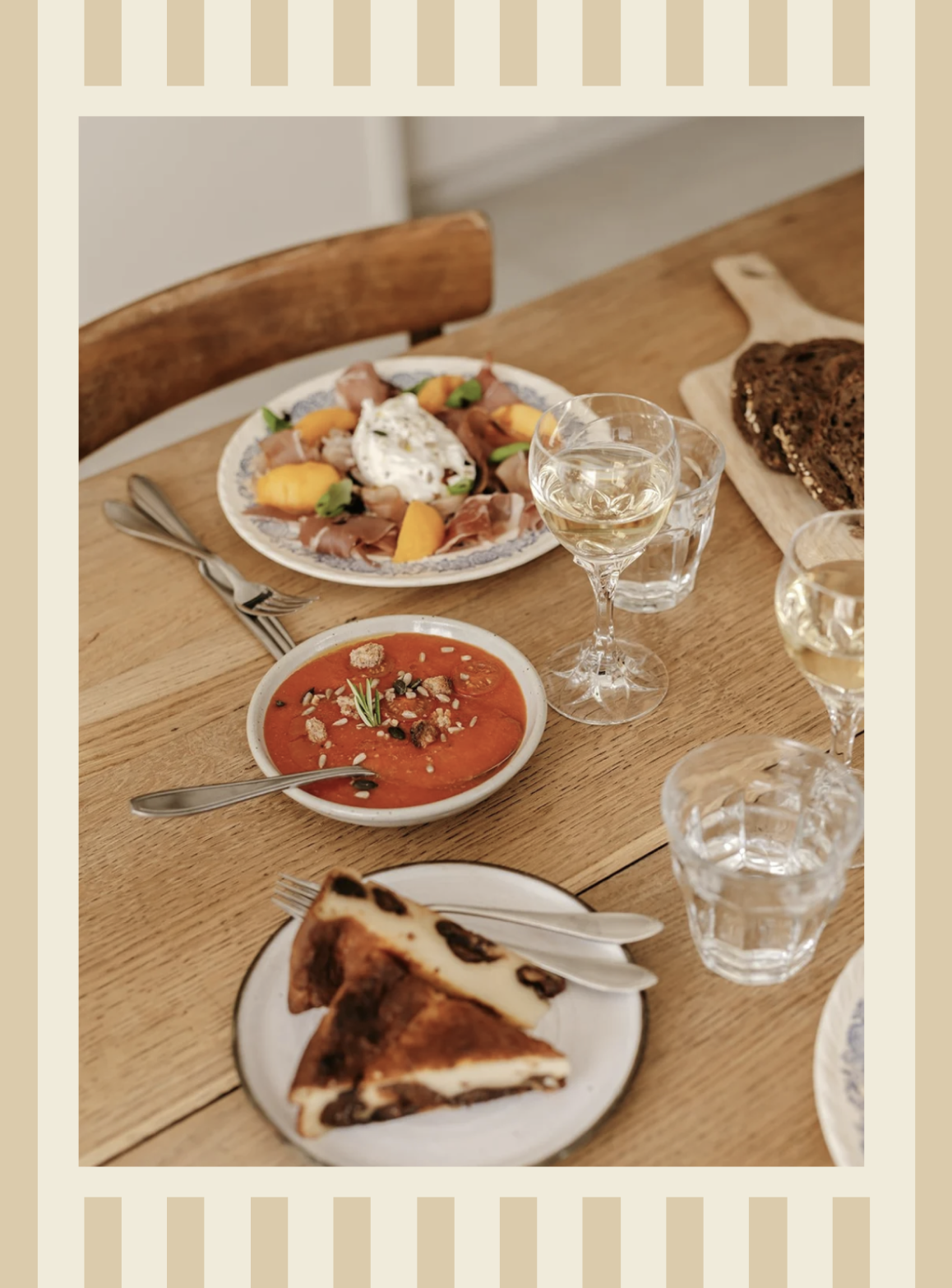 Repas avec une soupe à la tomate, une assiette de salade avec du fromage, un plat de pain complet, et deux verres de vin blanc sur une table en bois.