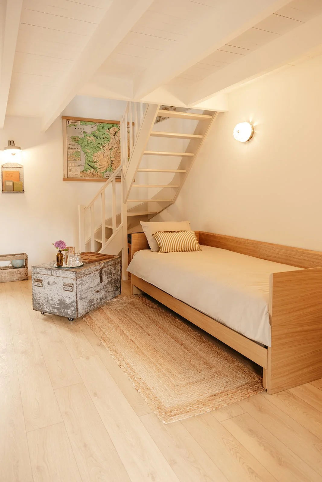 Une chambre cosy avec un lit en bois clair et une petite commode en métal repeinte en blanc, une étagère murale avec des livres et une lampe, une fresque géographique au mur, un tapis beige, et un escalier en bois menant à une mezzanine.