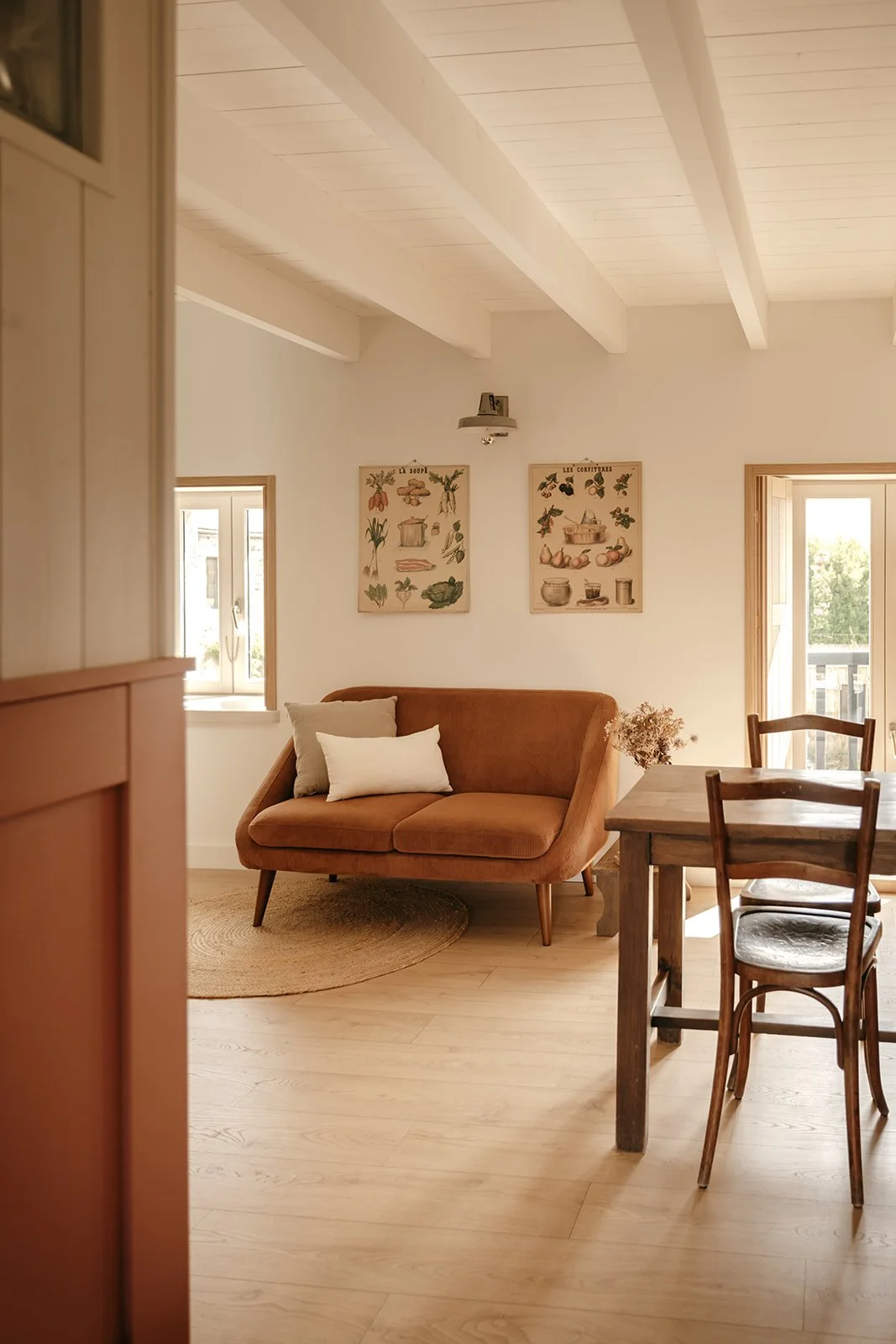 Salon cosy avec un canapé marron, un tapis rond, des chaises en bois, des planche scolaires vintage sur le mur, et des fenêtres laissant entrer la lumière naturelle.