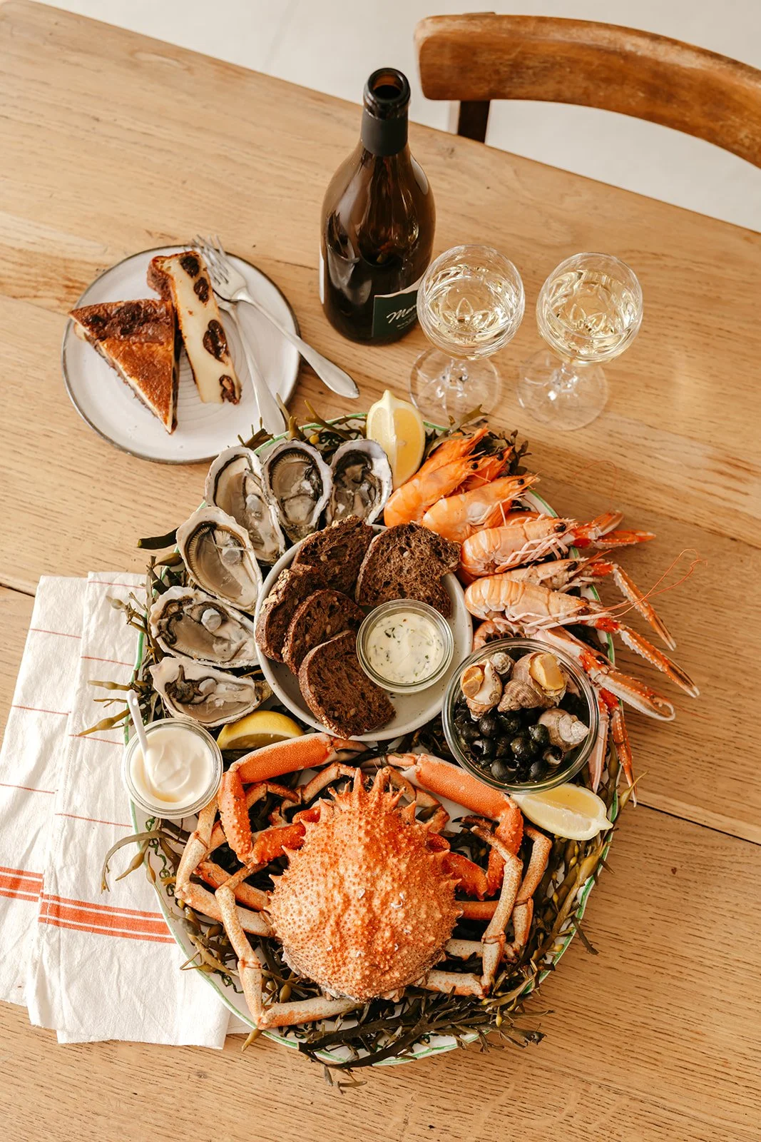 Plate de fruits de mer comprenant crabes, crevettes, huîtres, moules, et escargots, accompagnés de pain, beurre, et sauces, avec du vin et gâteau en dessert sur une table en bois.