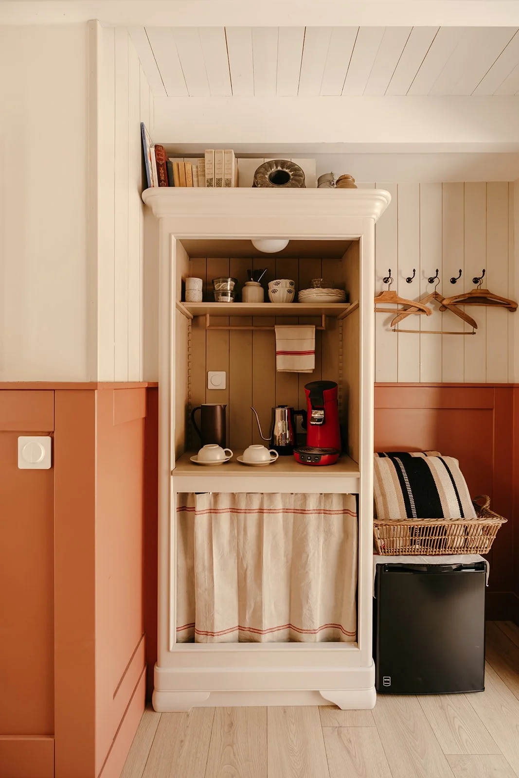 Un petit coin cuisine avec un ancien meuble transformé et contenant des tasses, une bouilloire, une cafetière électrique, une boîte de rangement, un torchon suspendu et un mini réfrigérateur