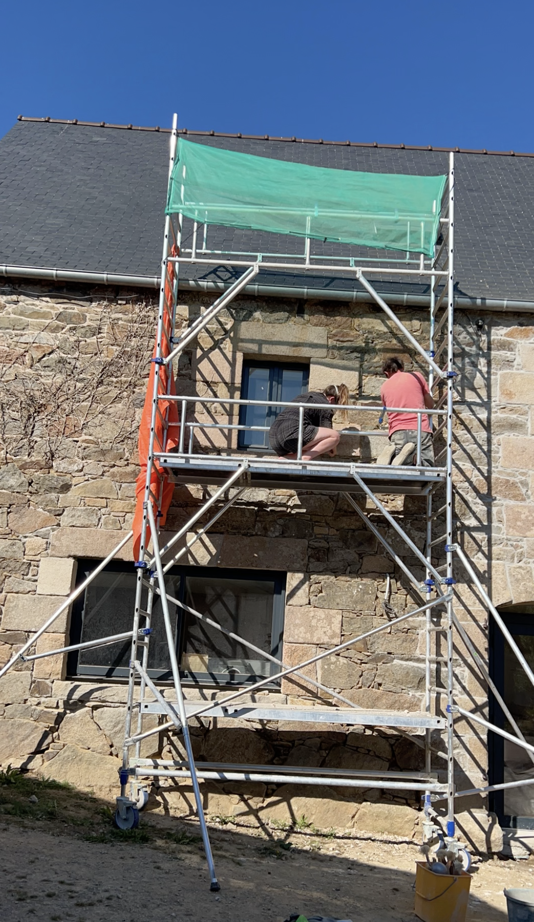 Deux personnes travaillent sur une échafaudage en face d'une maison en pierre, avec un ciel bleu en arrière-plan.