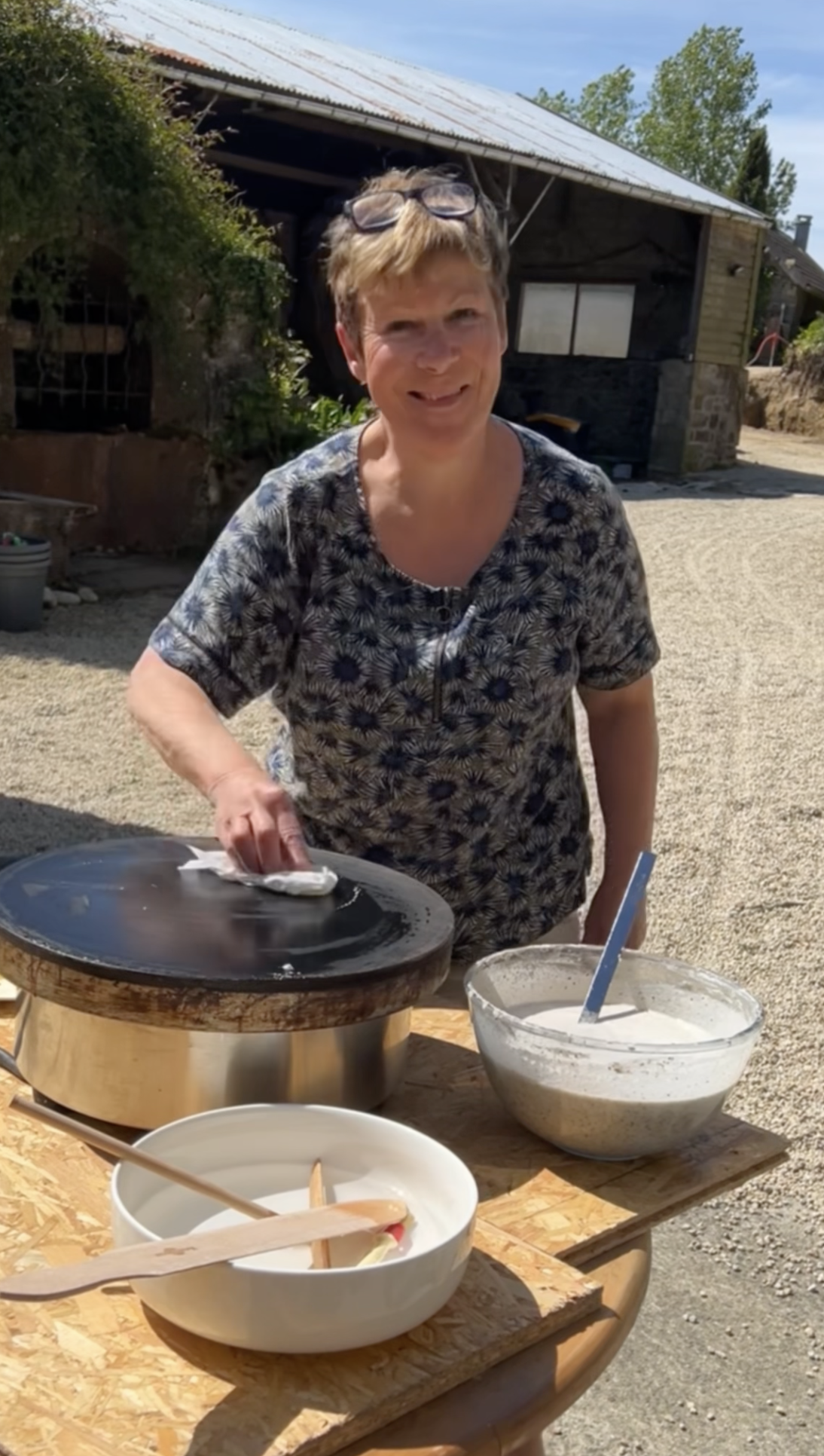 Une femme prépare une crêpe sur une bilié de la marque Krampouz en extérieur, avec des ingrédients comme de la pâte à crêpe dans un bol à côté.