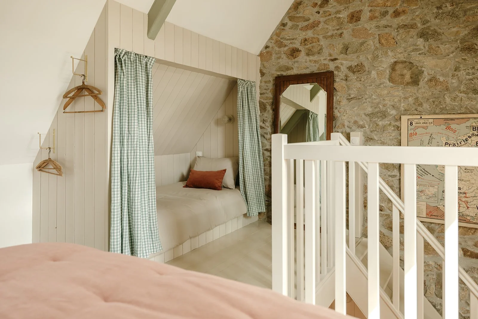 Une chambre avec un lit dans une niche sous pente, entouré de rideaux en vichy vert et blanc. Mur en pierre apparente, miroir vintage rectangulaire en bois et carte scolaire vintage accrochée au mur.