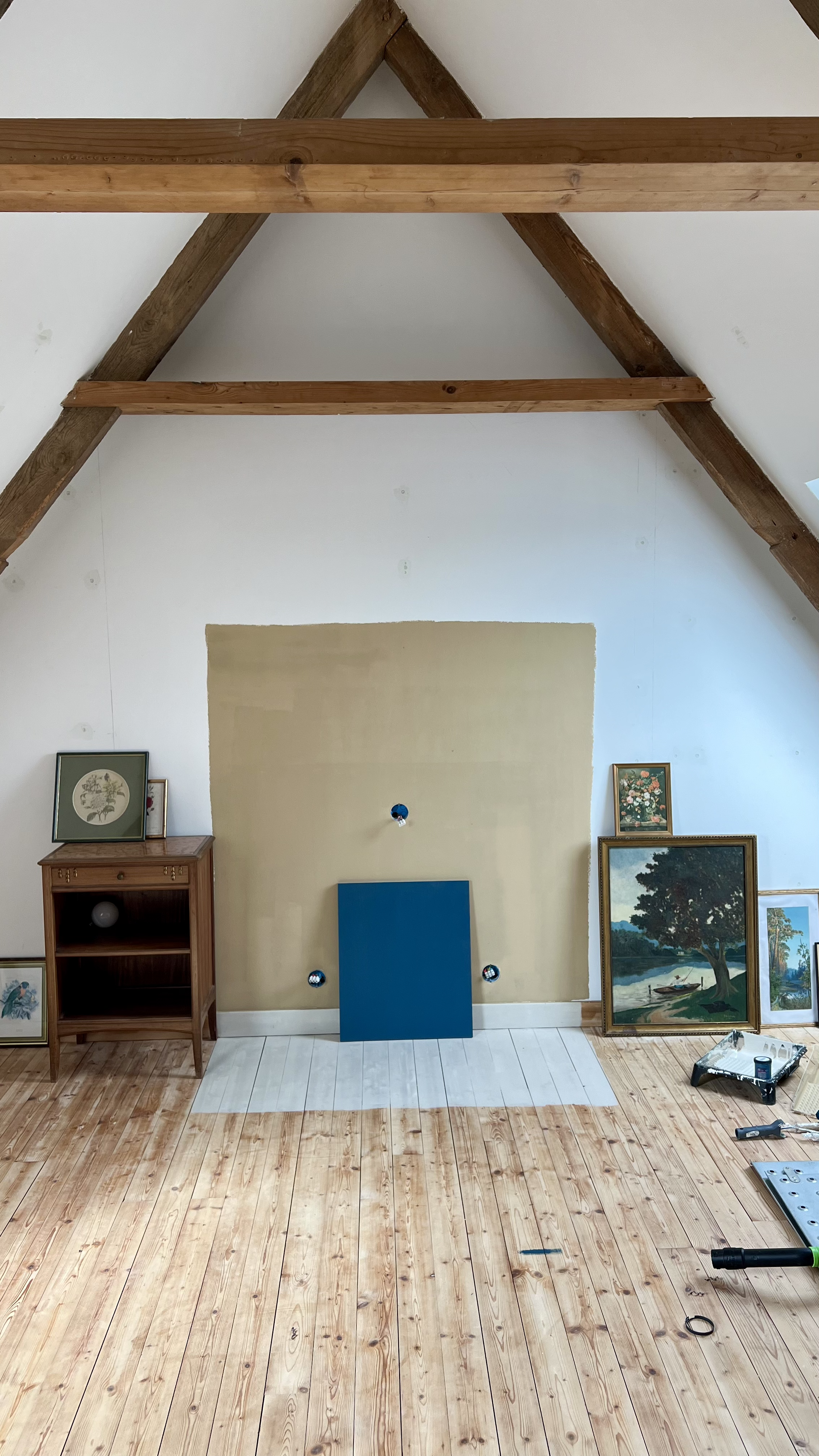 Une pièce d'intérieur en cours de rénovation avec un sol en parquet clair, plusieurs tableaux et cadres accrochés ou posés contre le mur, une étagère en bois, et des outils de bricolage.