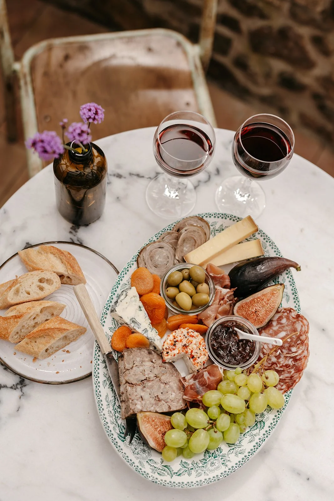 Plate de fromage, charcuterie, fruits, et pain, avec deux verres de vin rouge sur une table en marbre. Petites fleurs violettes dans un vase en verre.
