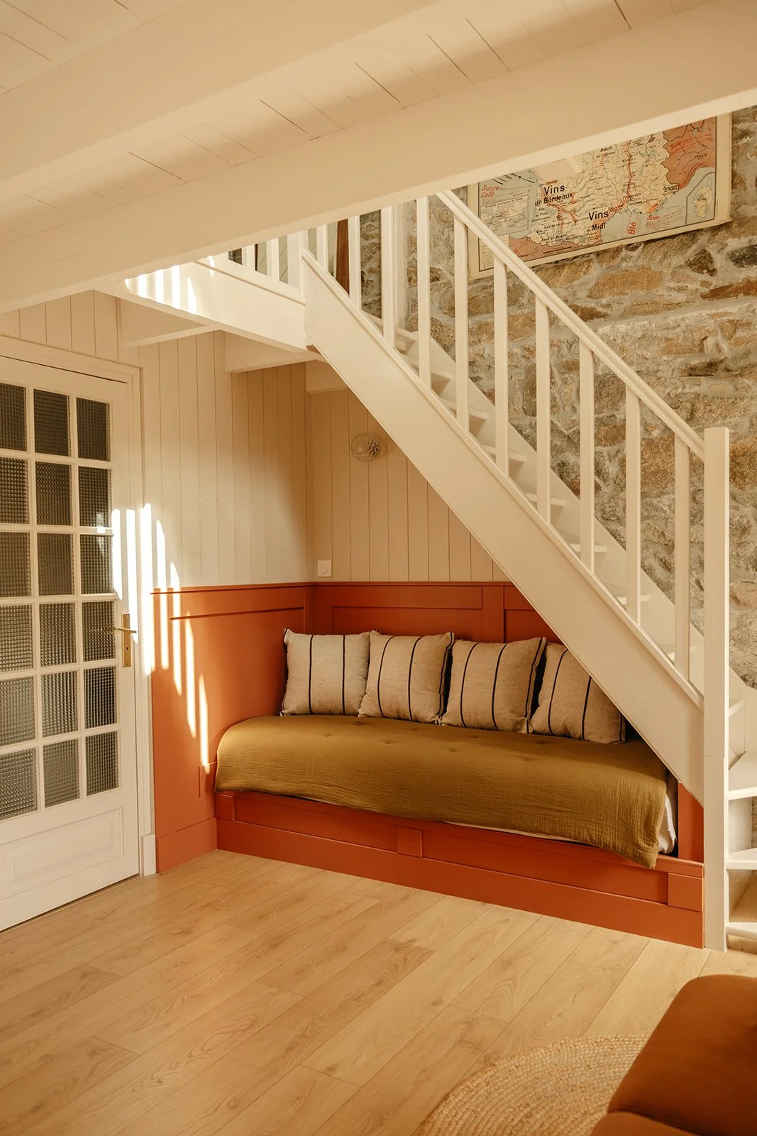 Intérieur de la chambre Dulse du Clos des Sentiles à Perros-Guirec avec une banquette en bois, des coussins, un escalier blanc en bois menant au premier étage, mur en pierre et carte scolaire vintage accrochée au mur.