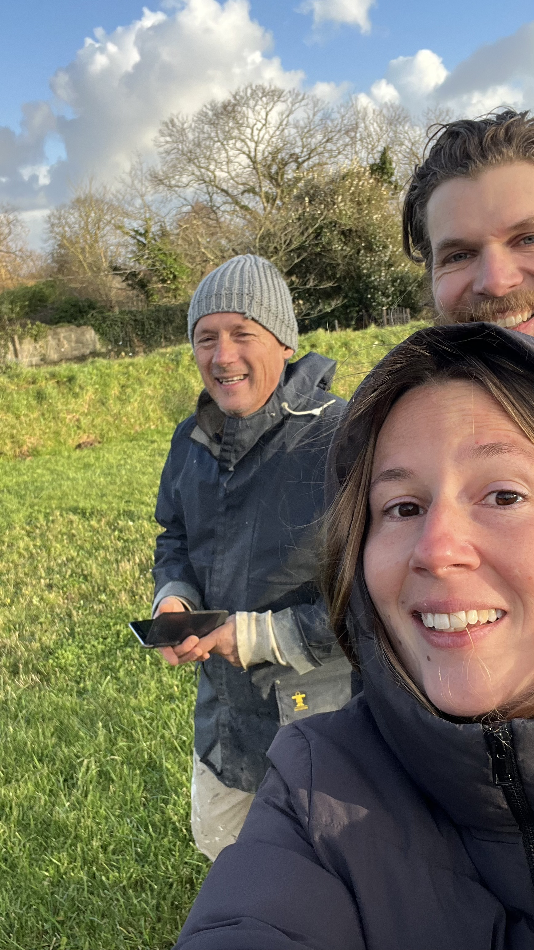 Trois personnes souriantes prennent une selfie en plein air avec un ciel nuageux et des arbres en arrière-plan.