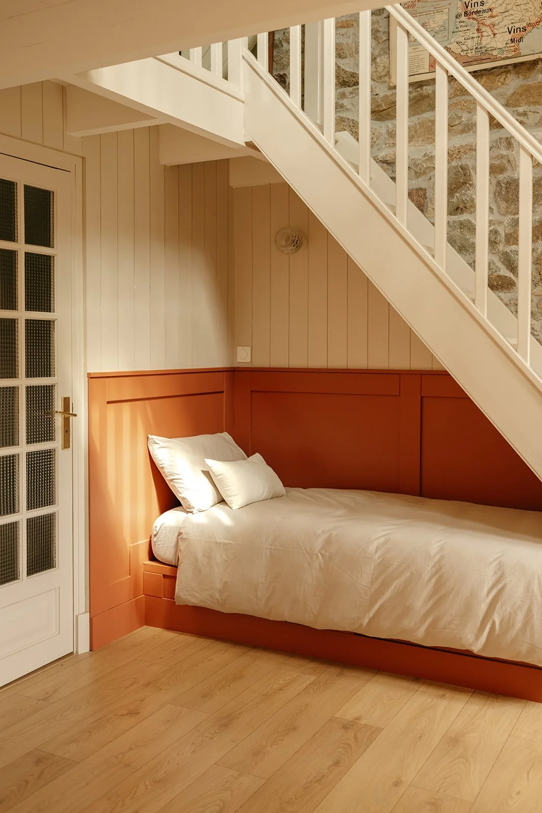 Lit simple sous un escalier dans une pièce chaleureuse avec mur en bois, fait face à une porte en verre avec cadre blanc.