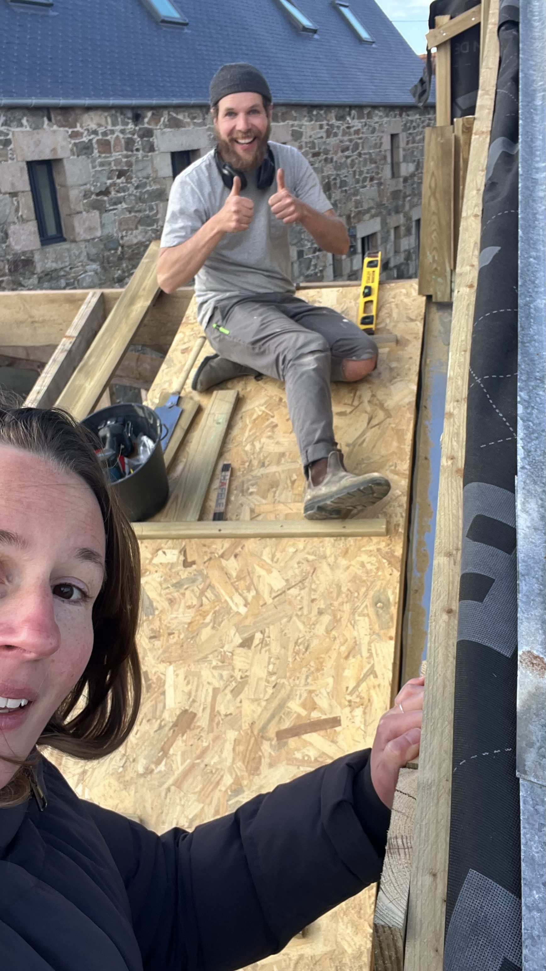 Deux personnes travaillant sur un chantier de construction en extérieur. Une femme prend une selfie avec un homme en arrière-plan qui sourit, donne un pouce en l'air et porte un casque de chantier. La structure en bois en construction est visible, ai