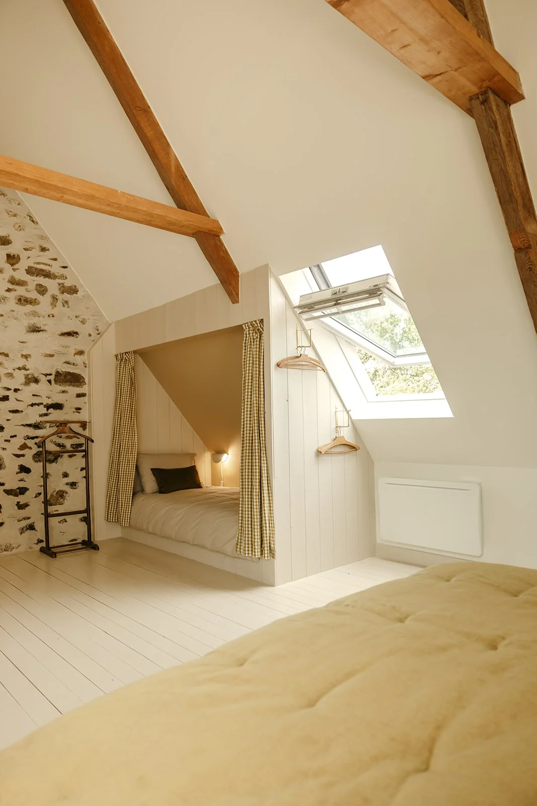 Chambre avec lit dans un espace sous pente, fenêtre de toit, mur en pierre et murs peints en blanc, avec accessoires comme une étagère à vêtements, des cintres en bois, et rideaux à carreaux jaunes et blancs.