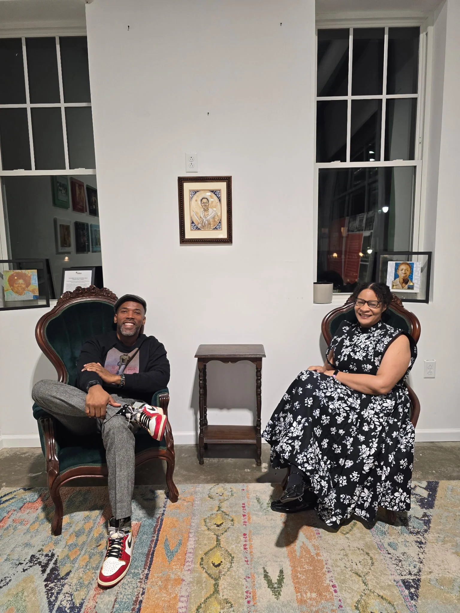 Two people seated in vintage chairs in an art gallery, smiling. A man with a beard wearing a black sweatshirt, gray pants, and colorful sneakers. A woman with glasses in a black and white floral dress. Behind them are five framed portraits on white walls and two large windows.