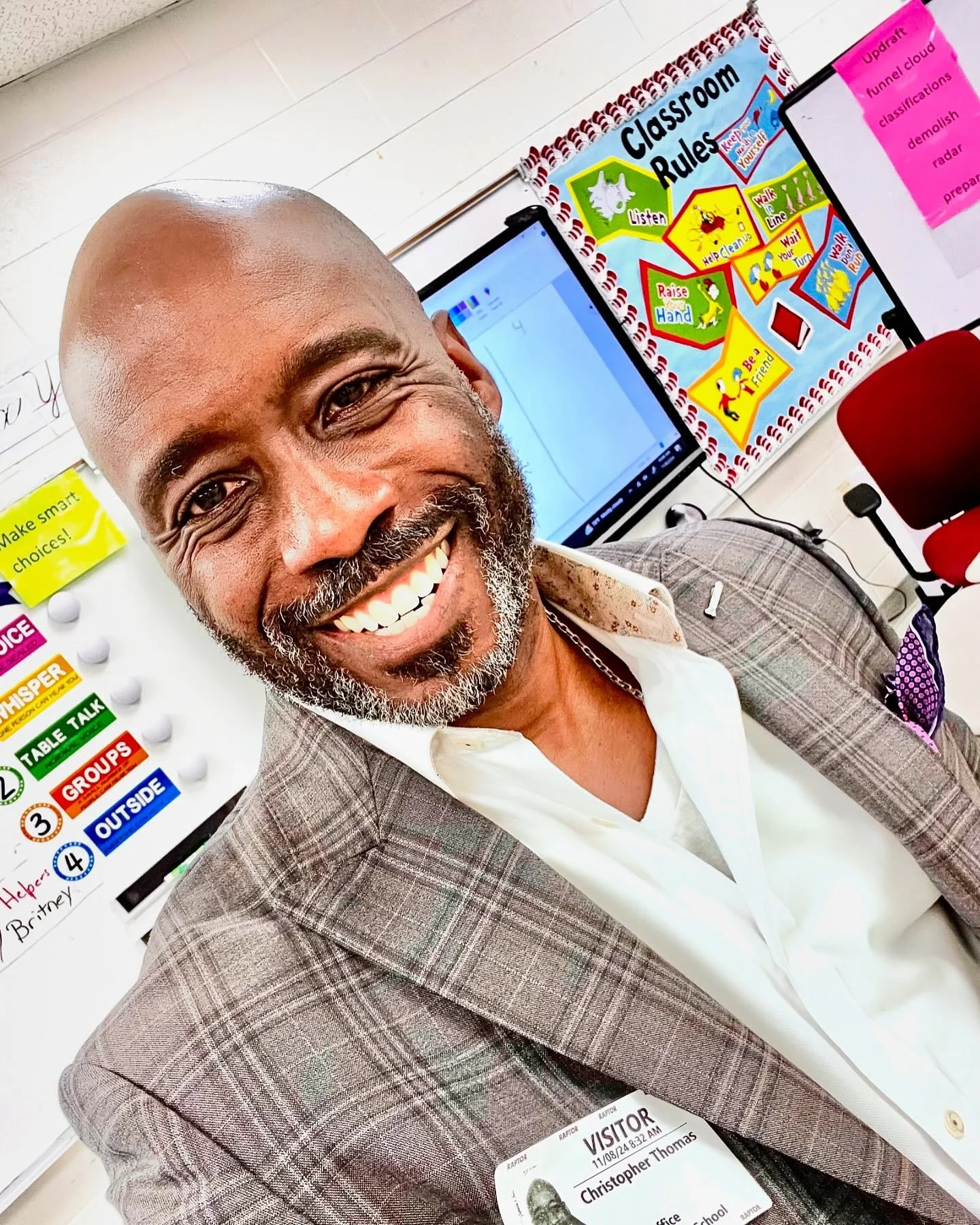 Smiling man with a beard and bald head wearing a plaid blazer and white shirt, standing in a classroom with colorful rules chart and computer screens in the background.