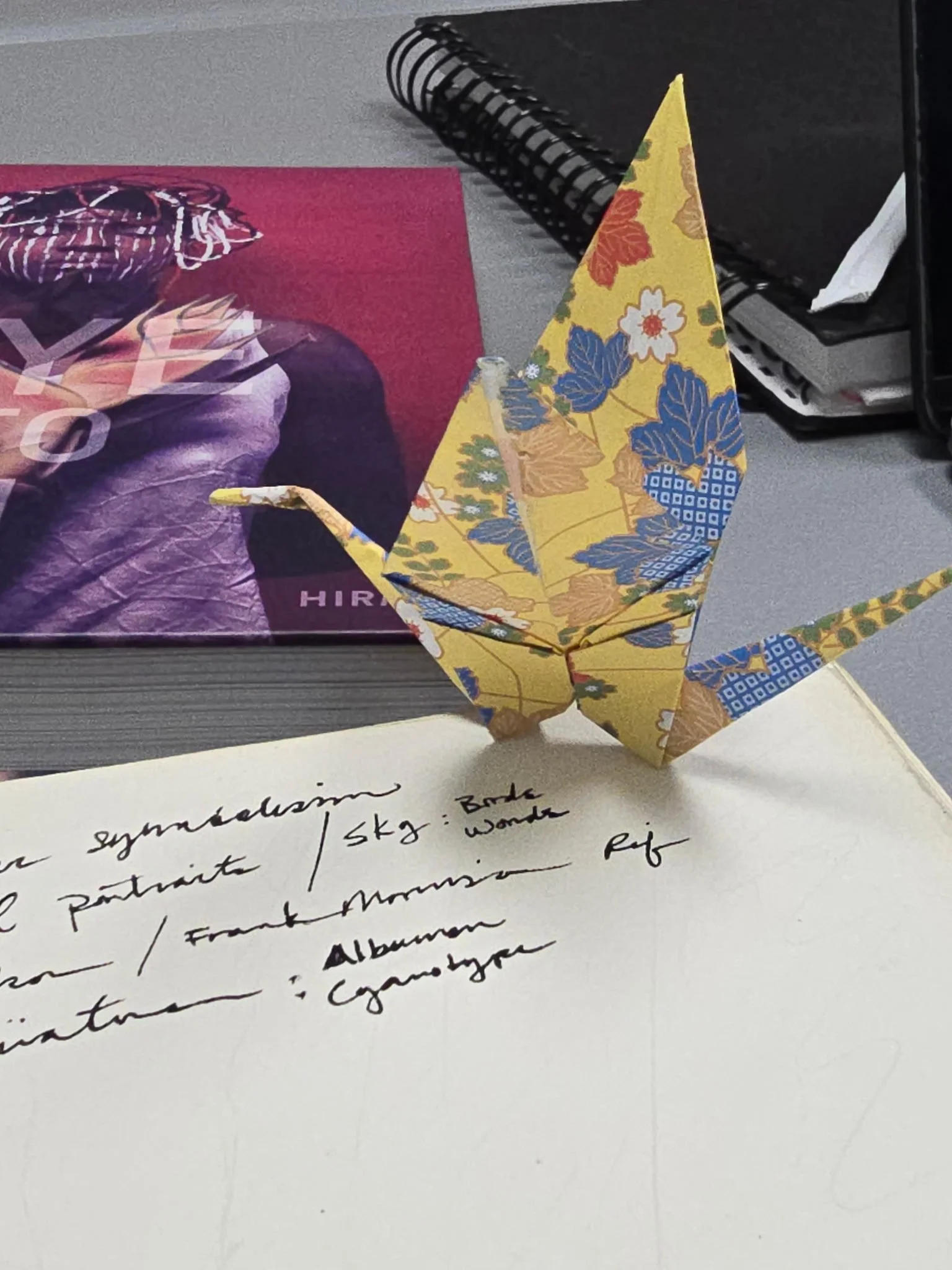 Colorful origami crane paper sculpture on a white surface, with handwritten notes underneath and office supplies in the background.