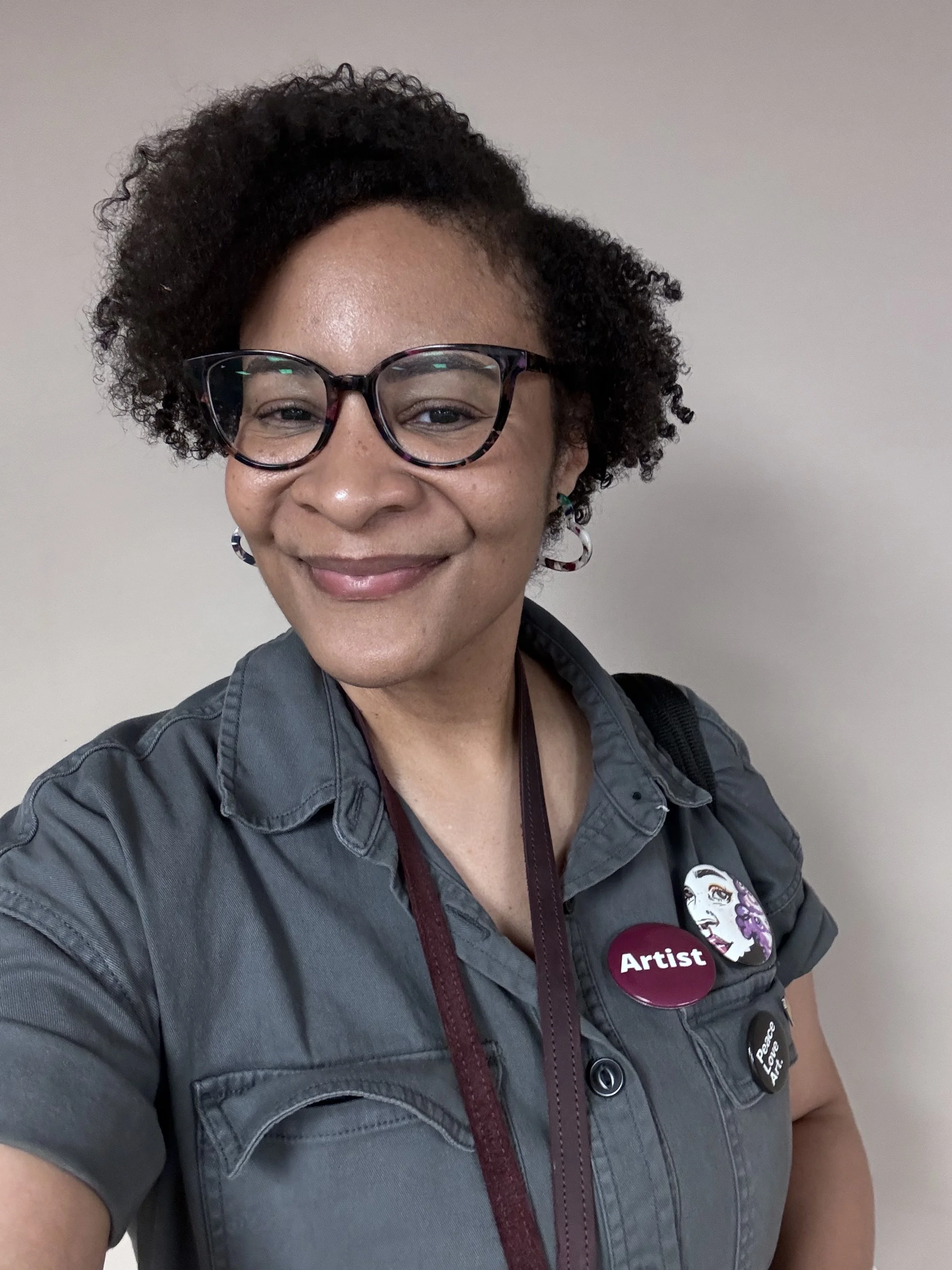 A woman with short curly hair wearing glasses, a gray shirt with decorative buttons, and badges that say "Artist" and "Peace Love Art." She has a slight shadow of facial freckles and is smiling softly against a plain light-colored background.