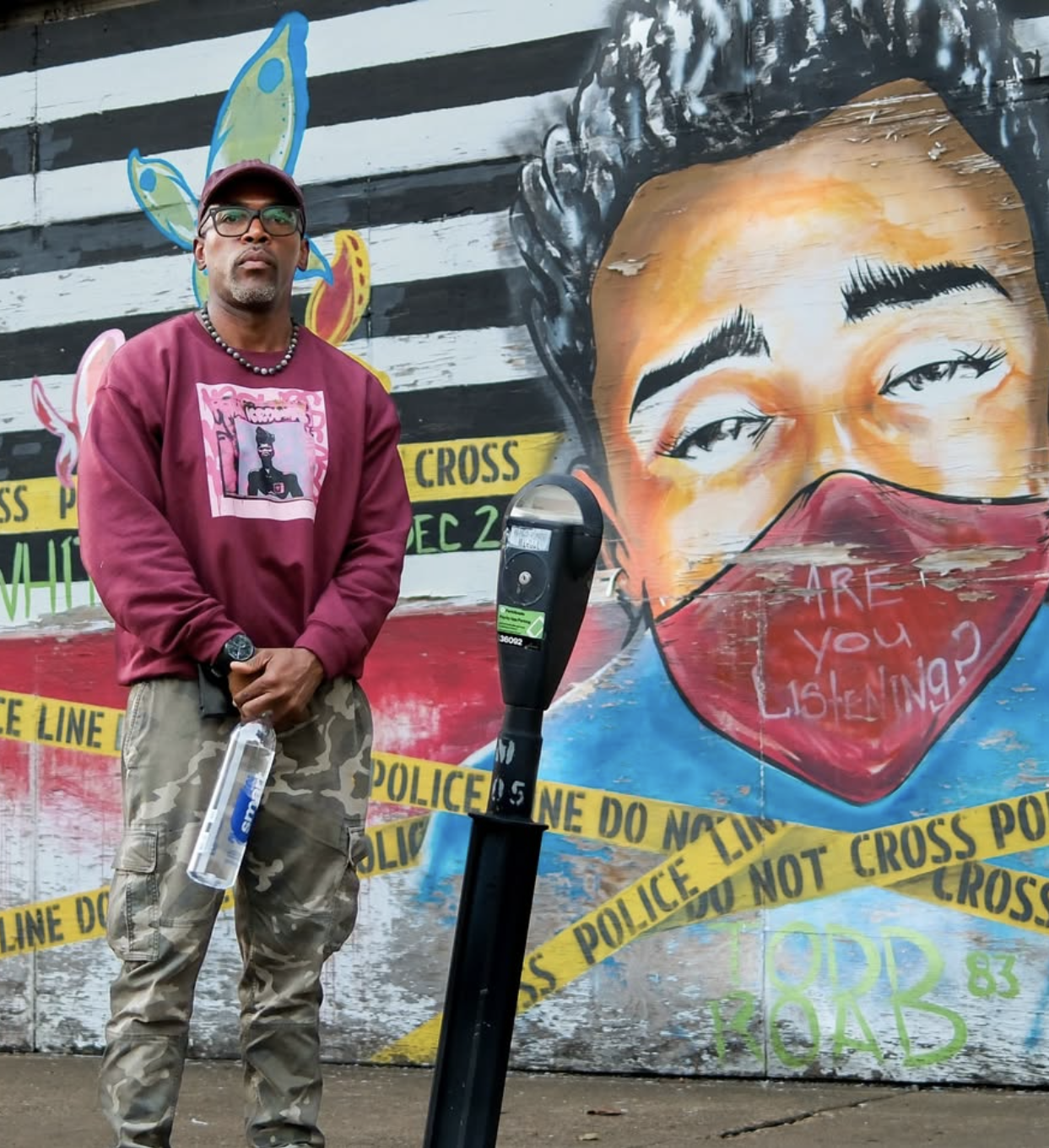 Man in a maroon sweatshirt and camouflage pants standing in front of a colorful mural depicting a woman's face with an orange face mask that has the words "ARE YOU LISTENING?" written on it. The mural is behind yellow police tape marked "POLICE LINE DO NOT CROSS." The man is holding a water bottle and standing near a parking meter.