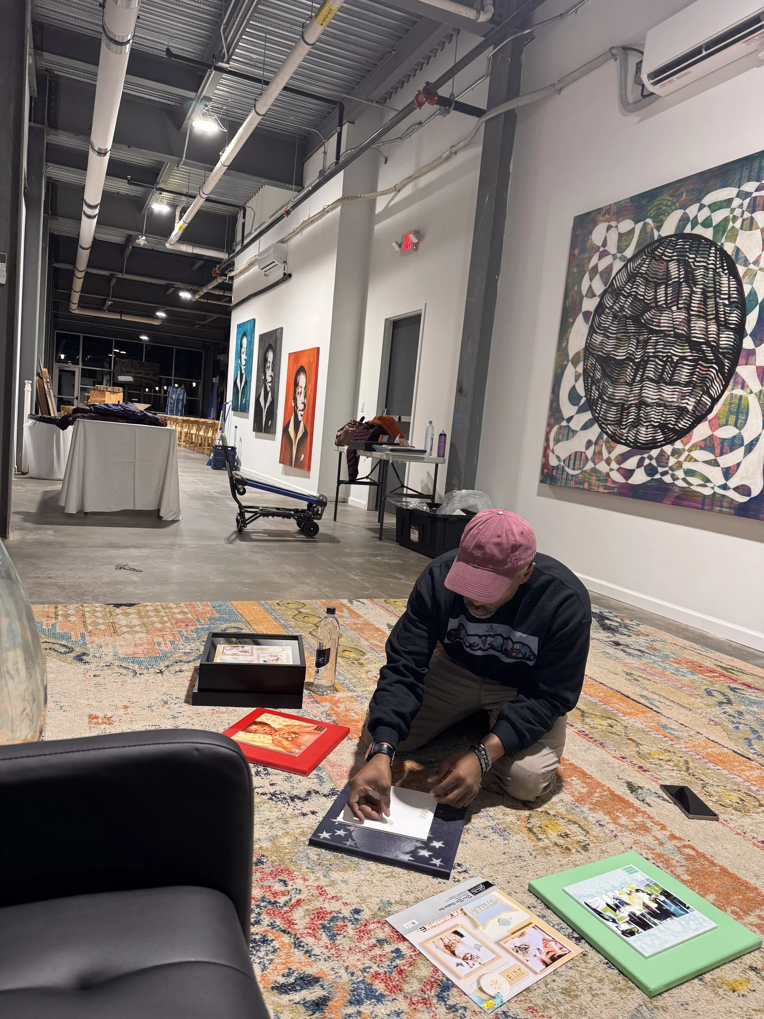 A man wearing a pink cap and black jacket is kneeling on the carpeted floor, engaging with various printed materials and picture frames on the ground, in an art gallery or exhibition space. Background features white walls with colorful artwork, tables with items, and a man sitting on a black leather sofa.