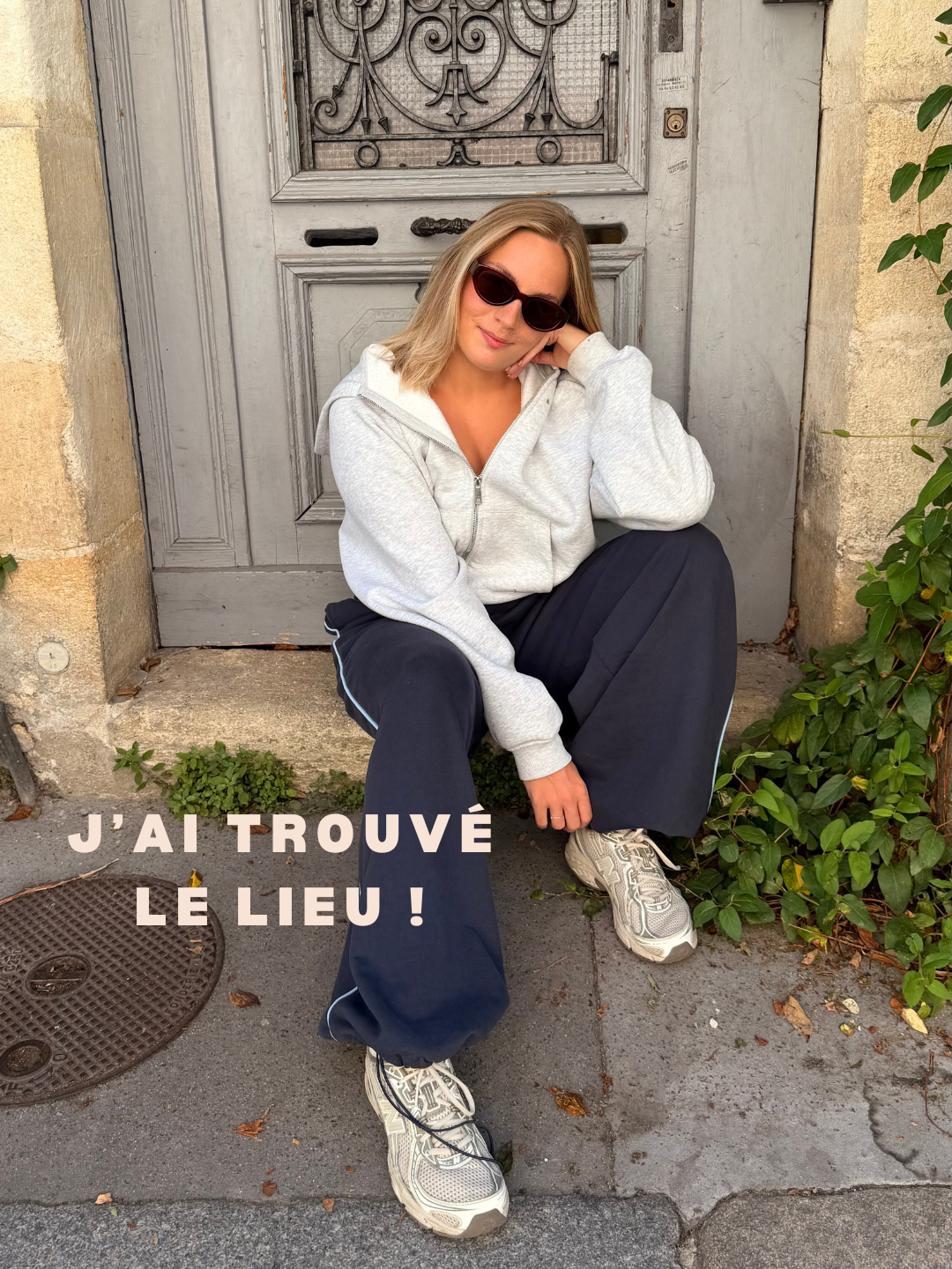 Une femme assise devant une porte en bois grise, portant des lunettes de soleil, un sweat gris, un pantalon noir et des baskets blanches. Texte en français sur l'image : « J'ai trouvé le lieu ! »