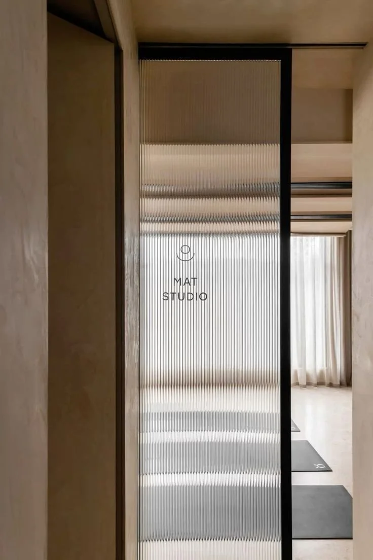 Une porte en matériau translucide avec l'inscription 'MAT STUDIO', située dans un intérieur modern. À l'arrière-plan, on voit une pièce avec des rideaux blancs et un tapis de pilates.