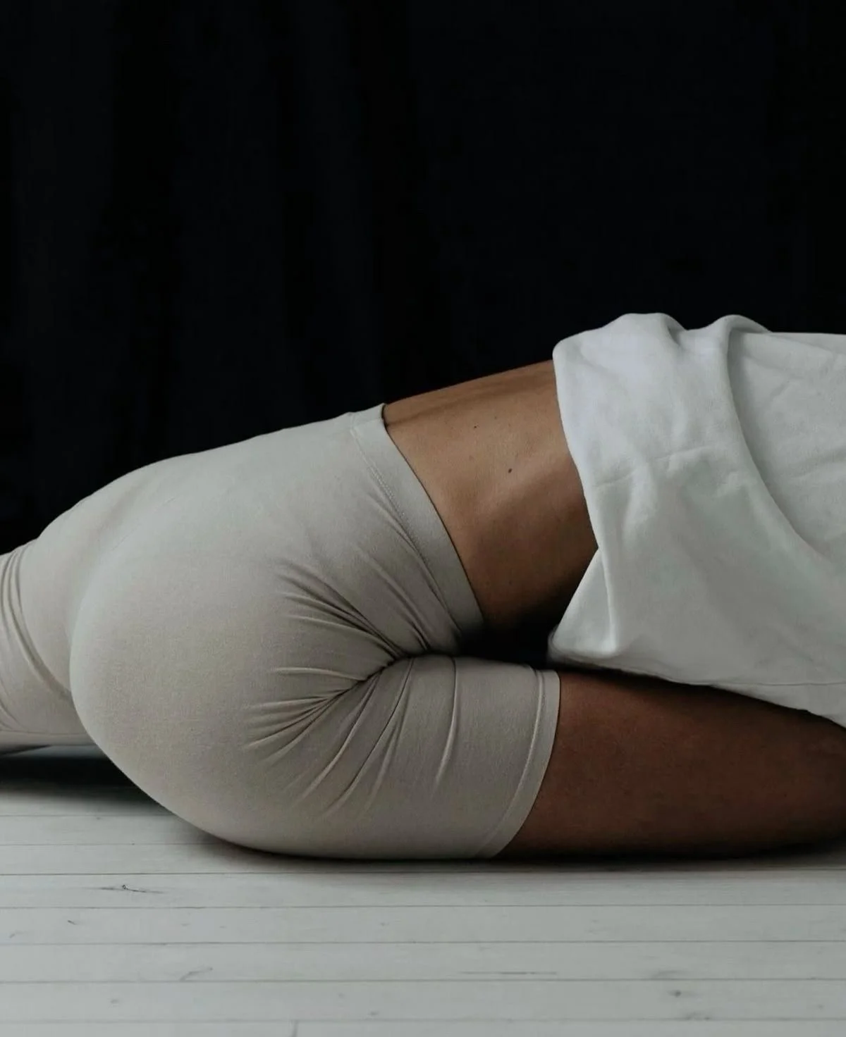 Une personne allongée sur le sol, portant un short beige et un t-shirt blanc, avec une toile blanche sur le haut du corps, contre un fond noir.