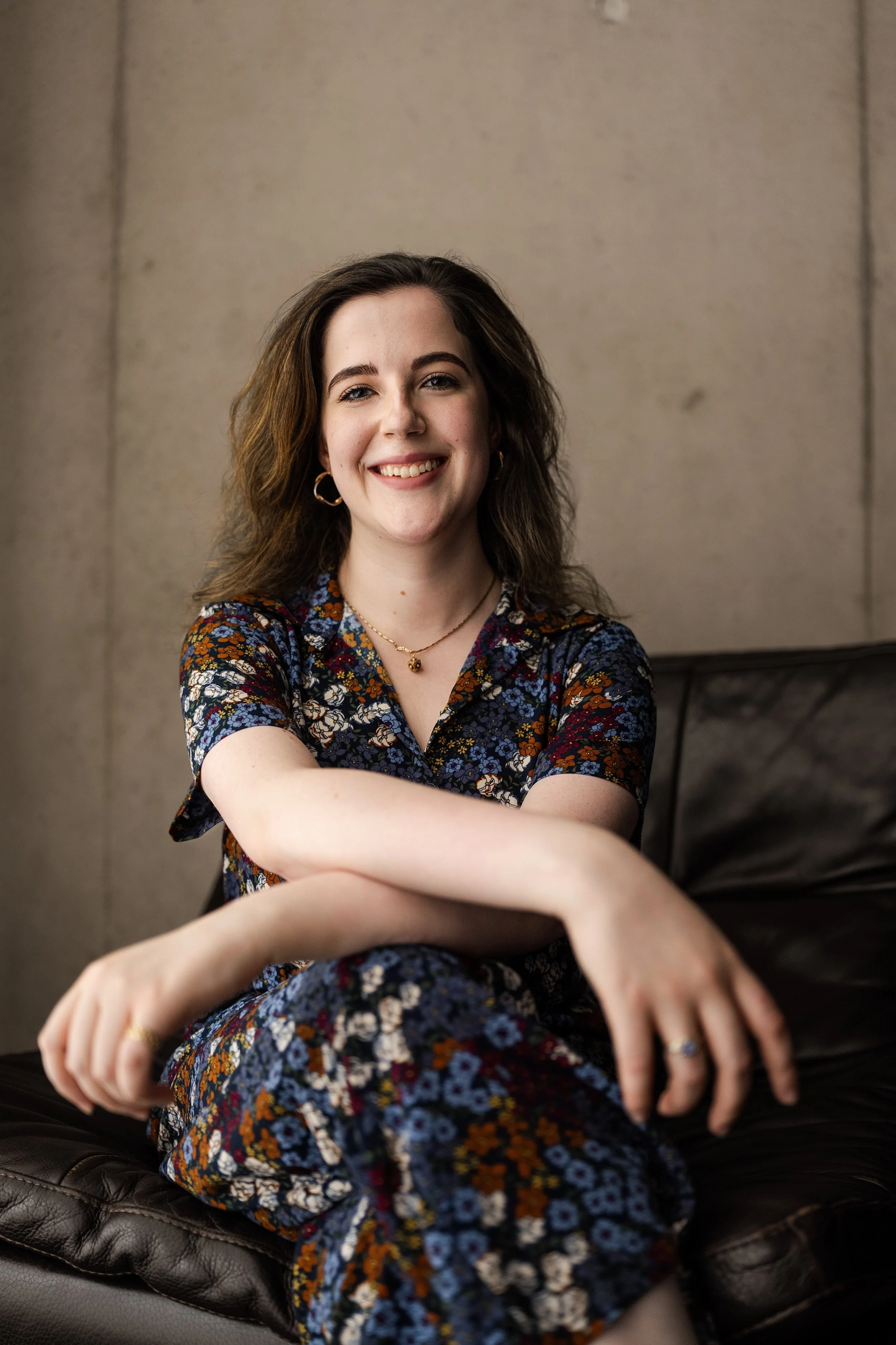 Portrait of a young woman with wavy brown hair, smiling, wearing gold hoop earrings, a floral dress, and gold necklace, sitting on a leather couch against a plain beige wall.