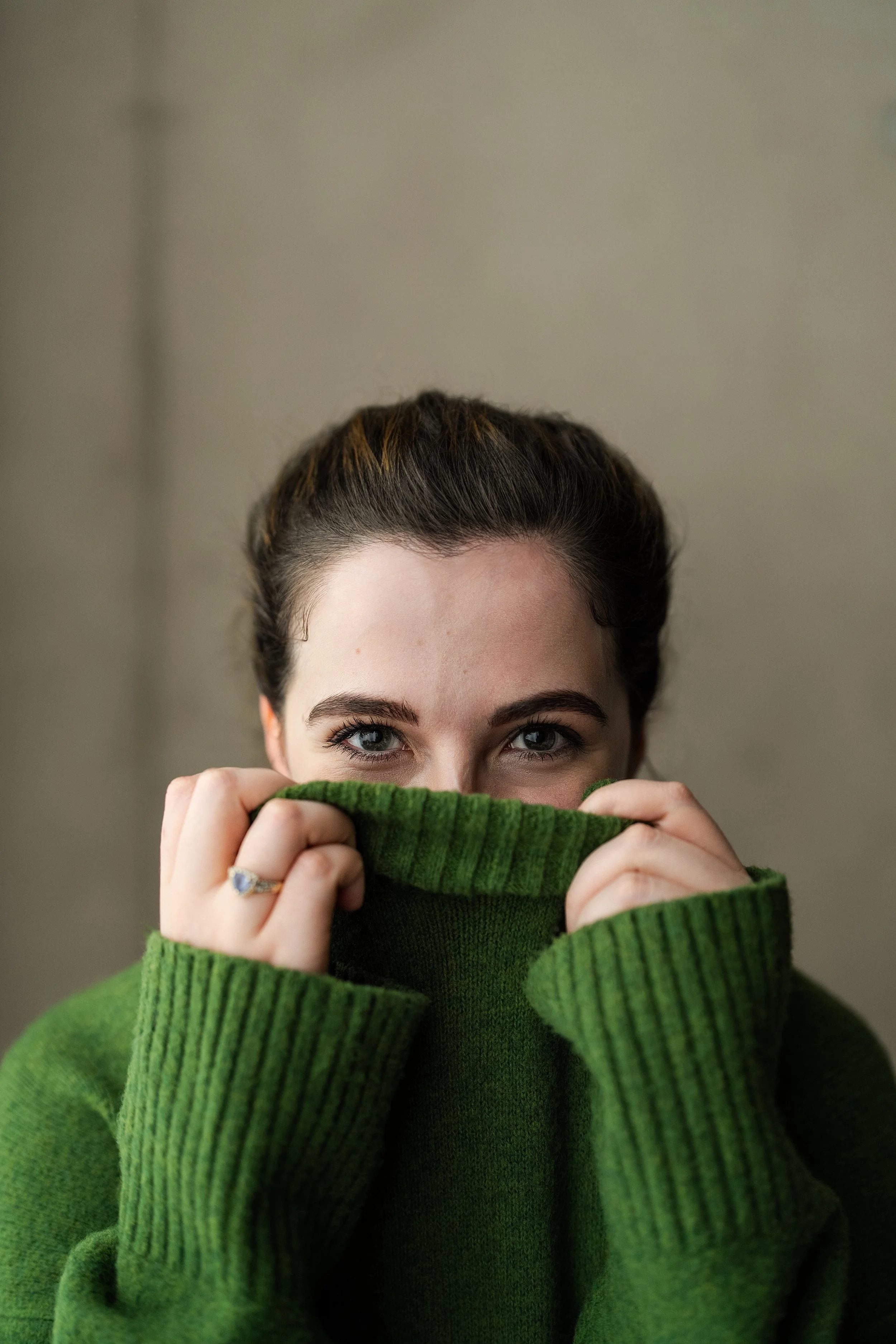 A woman with blue eyes and dark brown hair pulled back, wearing a green sweater and holding it up to partially cover her face.