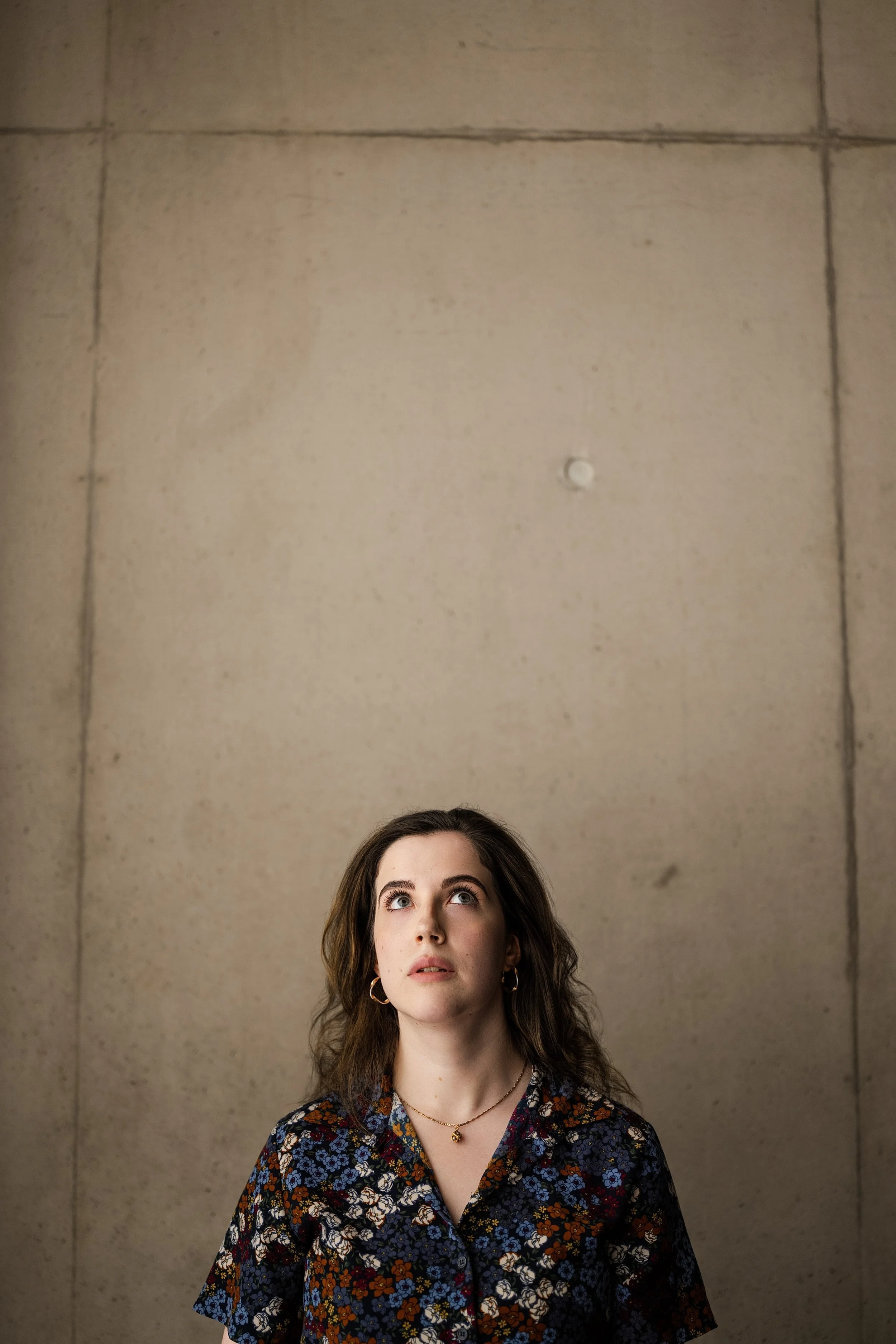 A young woman with long wavy hair looking up, standing in front of a plain concrete wall. She's wearing a floral patterned blouse and gold hoop earrings.