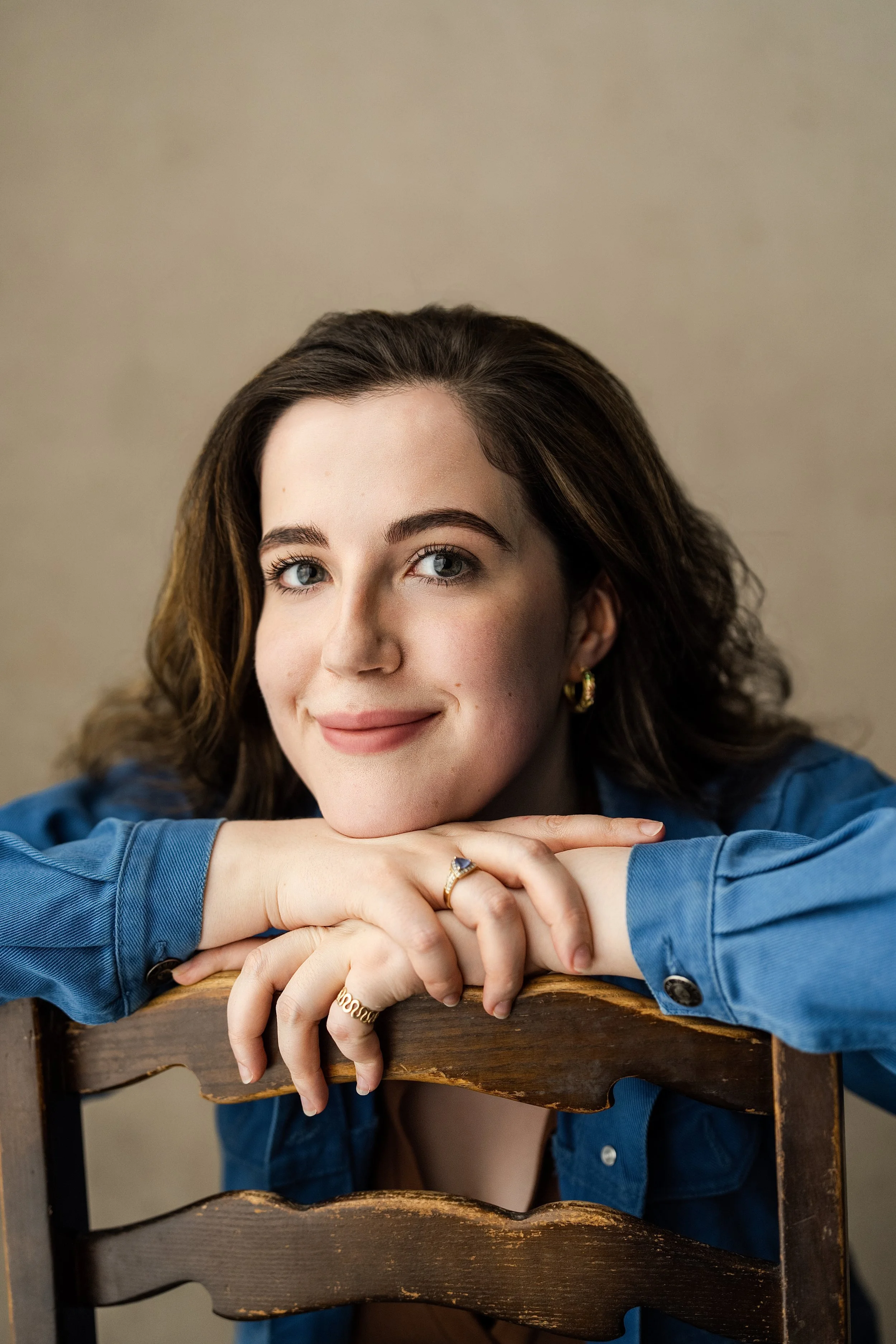 Close-up of a young woman with brown hair, blue eyes, and a light smile, resting her chin on her folded arms, wearing a blue shirt and gold jewelry, sitting behind a rustic wooden chair.