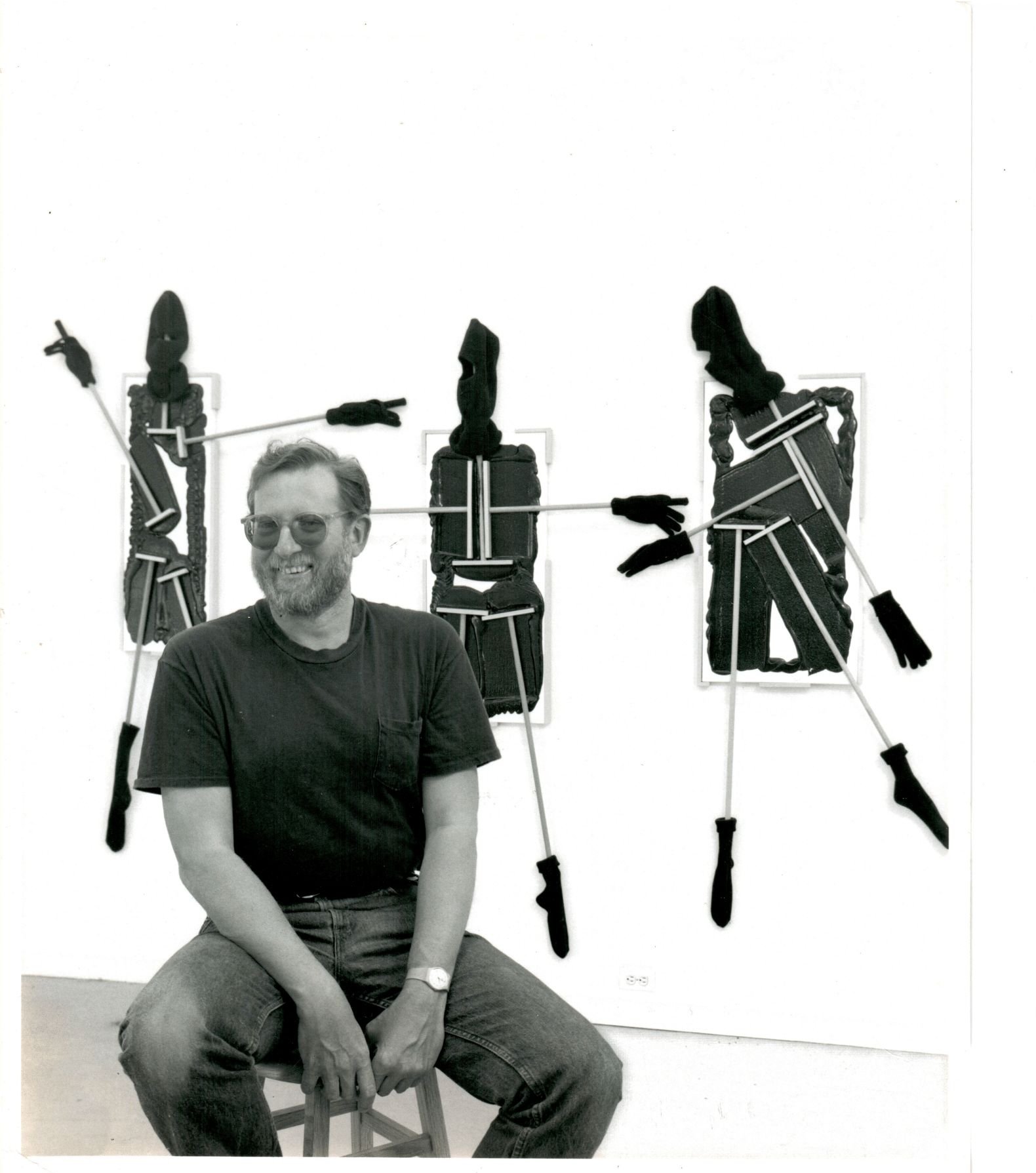 Black and white photo of a smiling man with glasses, sitting on a wooden chair, wearing a dark t-shirt and jeans, with three mannequin arms hanging on a wall behind him.